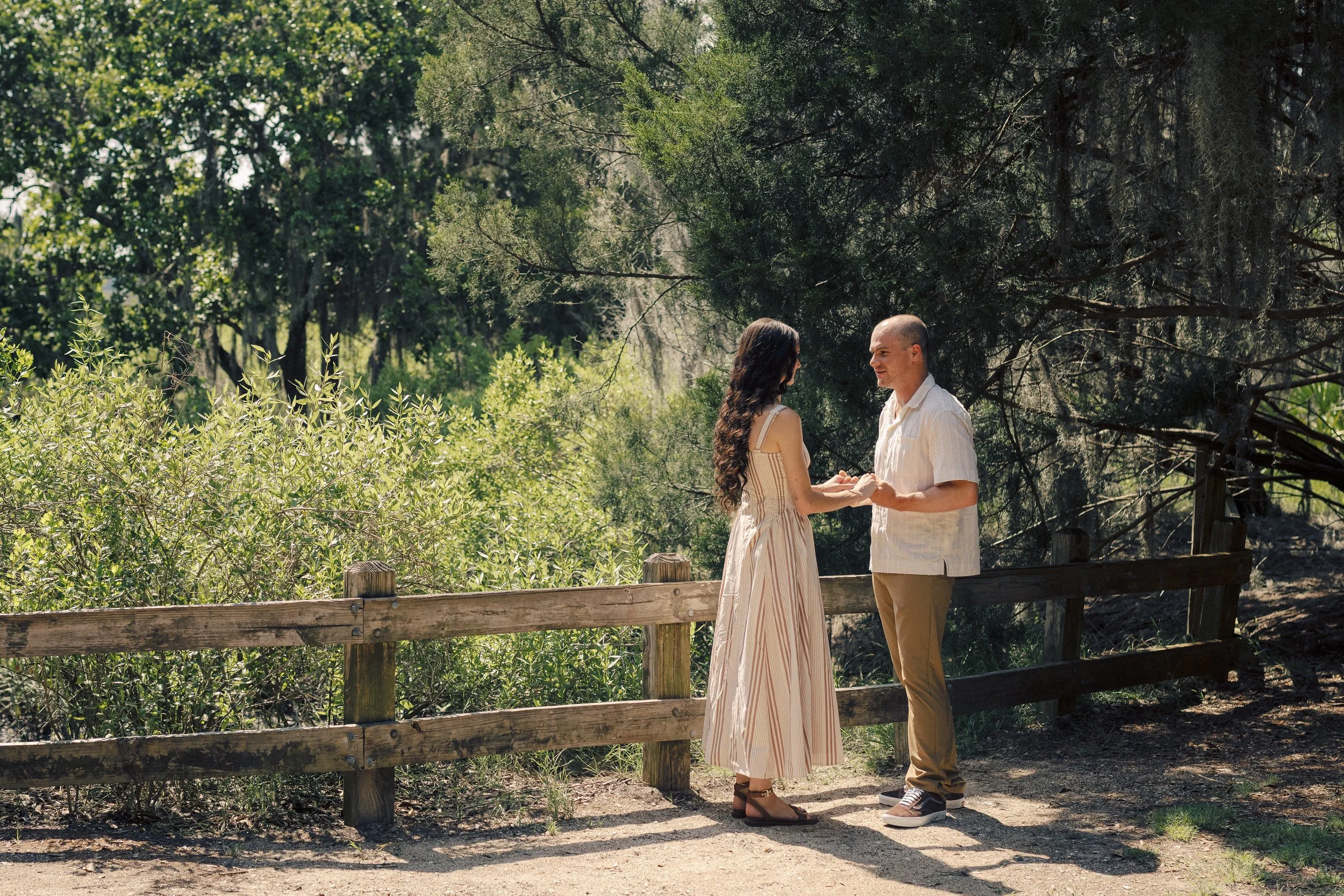 A man and woman holding hands and gazing at each other in a wooded outdoor setting during daytime.