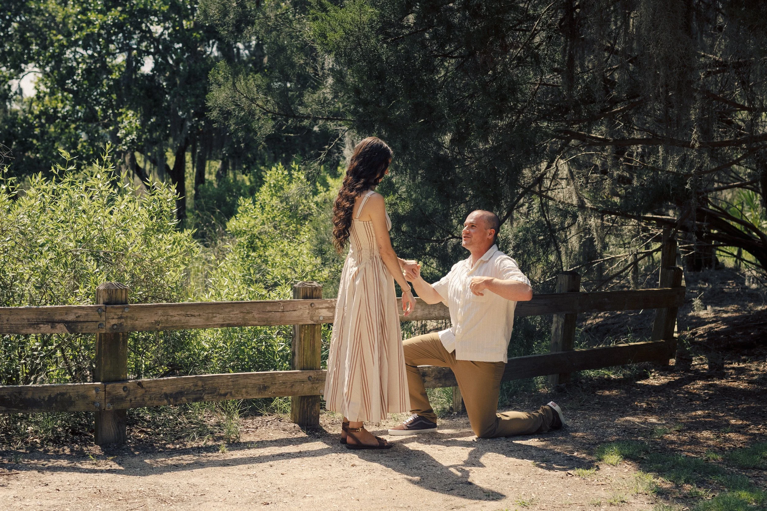 A man down on one knee proposing to a woman, who is standing in a park surrounded by trees and greenery, with a wooden fence in the foreground.
