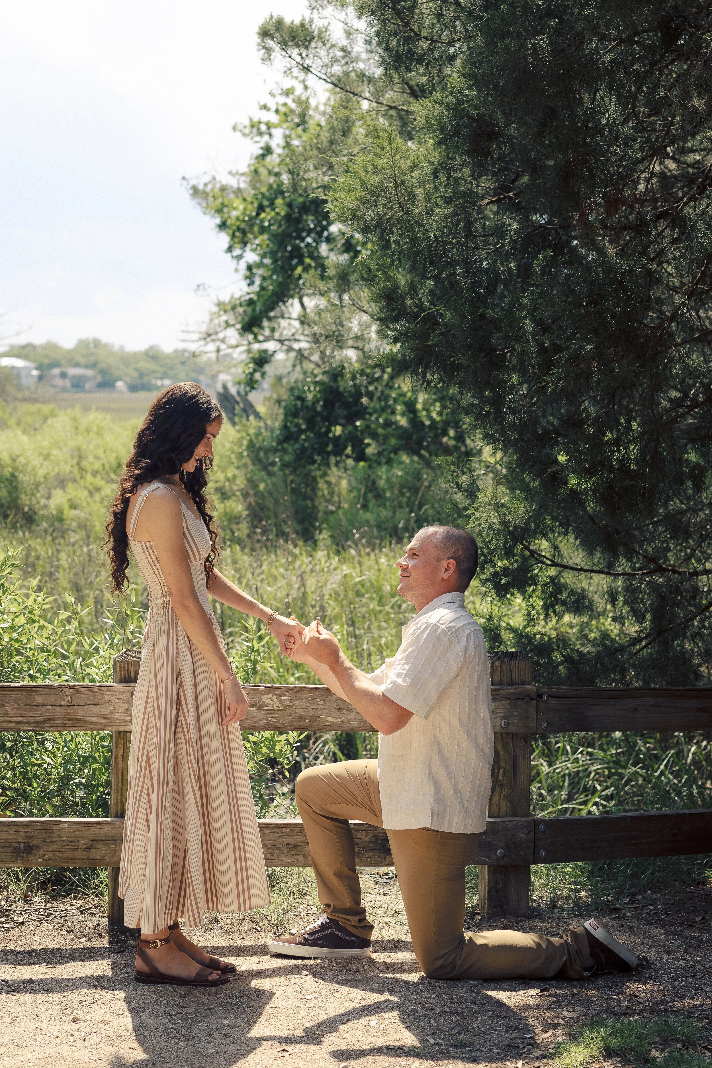 A man down on one knee, proposing to a woman in a park during daytime, holding her hands while she looks down at him. The woman has long dark wavy hair and is wearing a flowing striped dress and sandals. The man is dressed in a light-colored shirt an