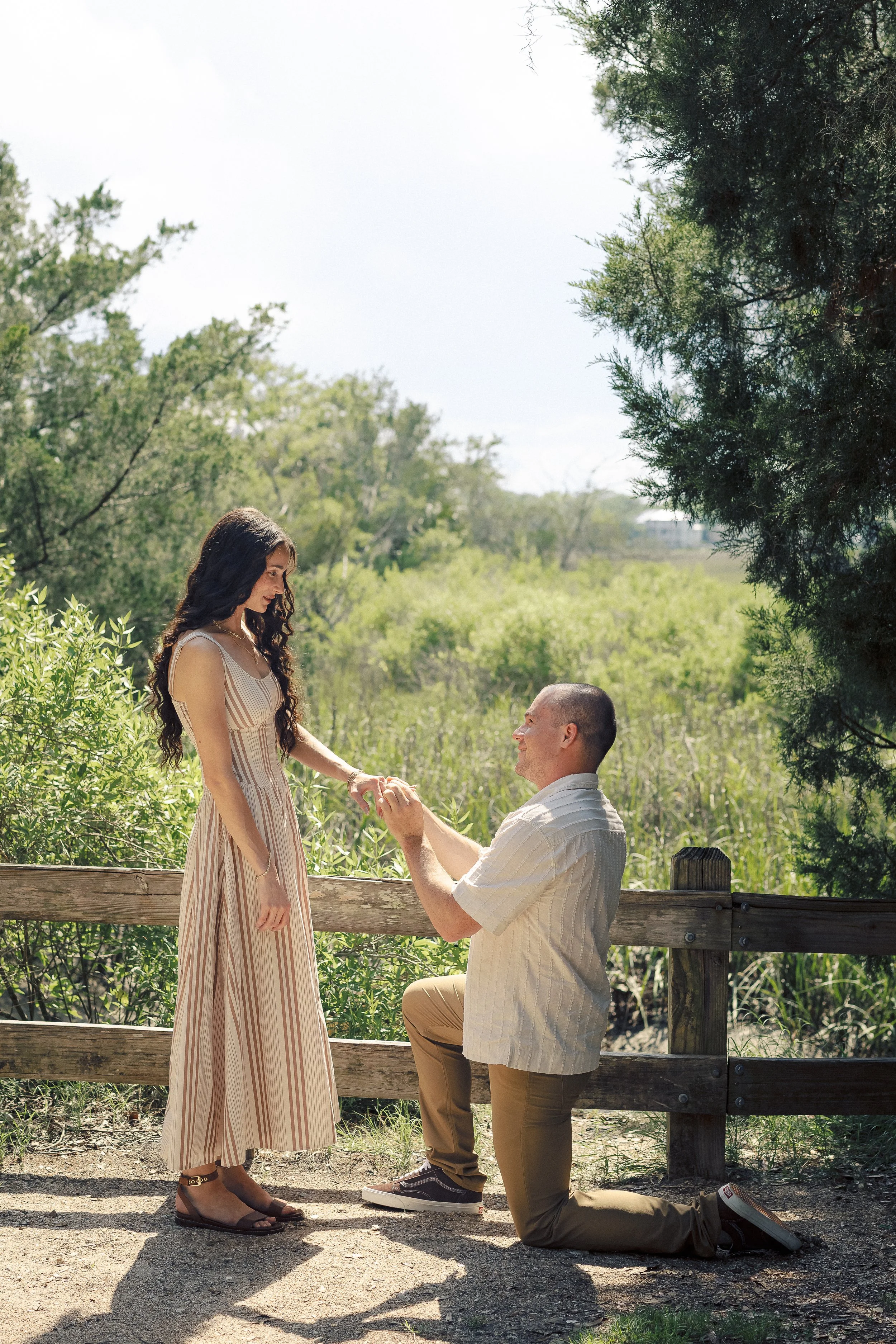 A man proposes to a woman outdoors on a wooden bridge, with the man kneeling and holding the woman's hand as she looks at him. The scene is set in a scenic, sunny area with trees and greenery.