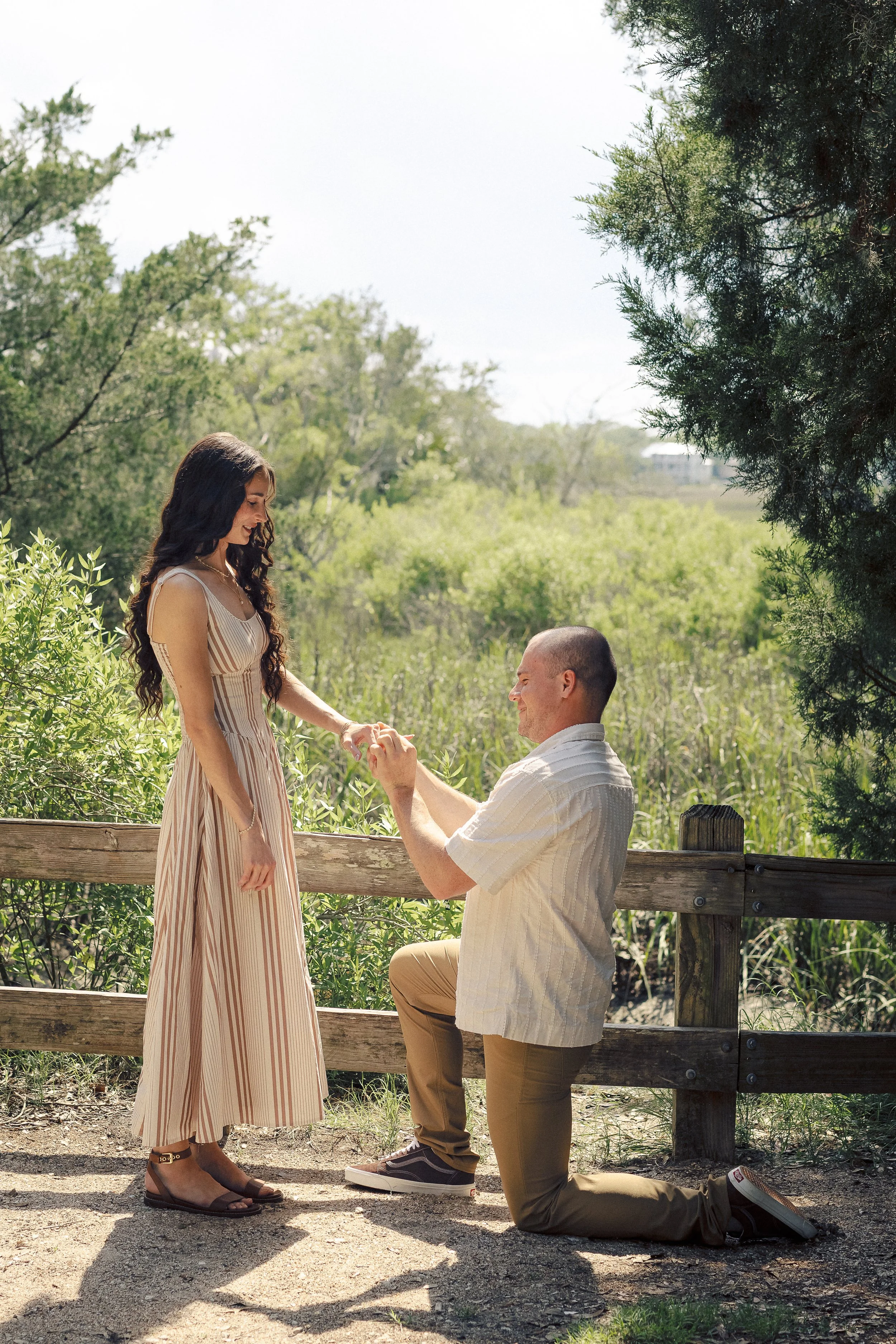 A man is kneeling on one knee, proposing to a woman on a walk outdoors in a green, wooded area. The woman is standing, holding the man's hand, and smiling. They are behind a wooden fence, with trees and a bright sky in the background.