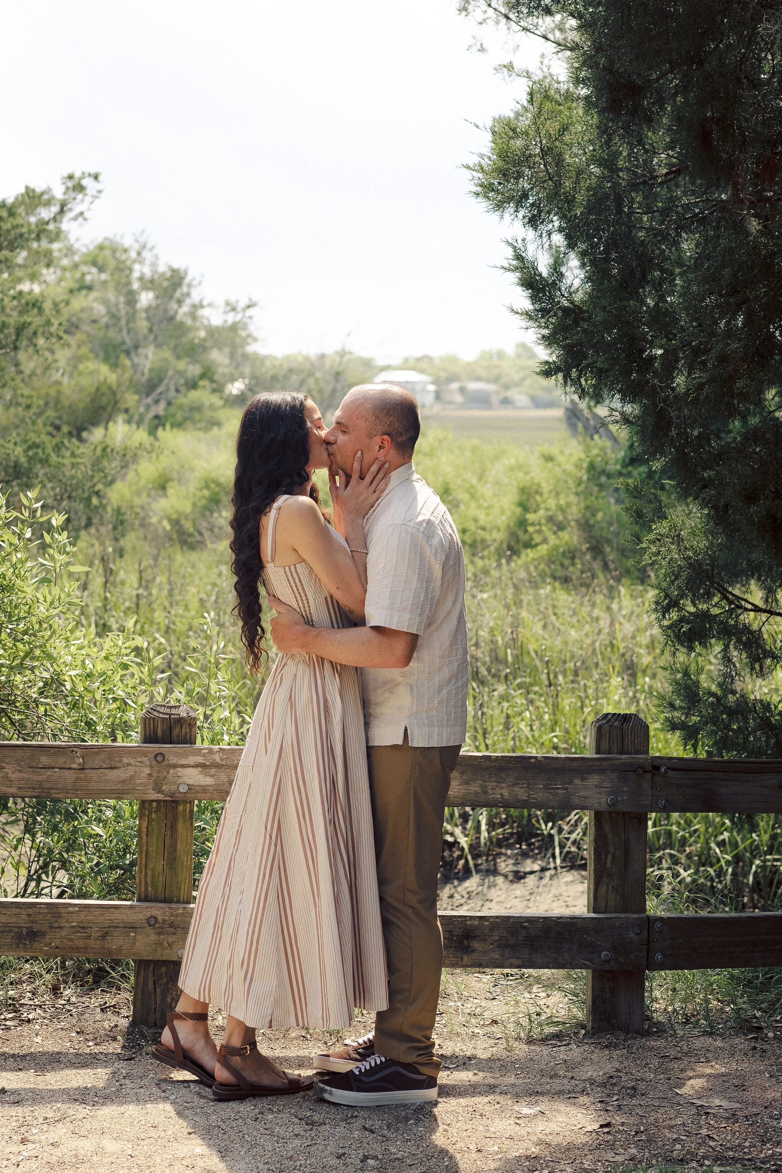 A couple kissing outdoors on a wooden bridge surrounded by green trees and bushes, with a clear sky in the background.