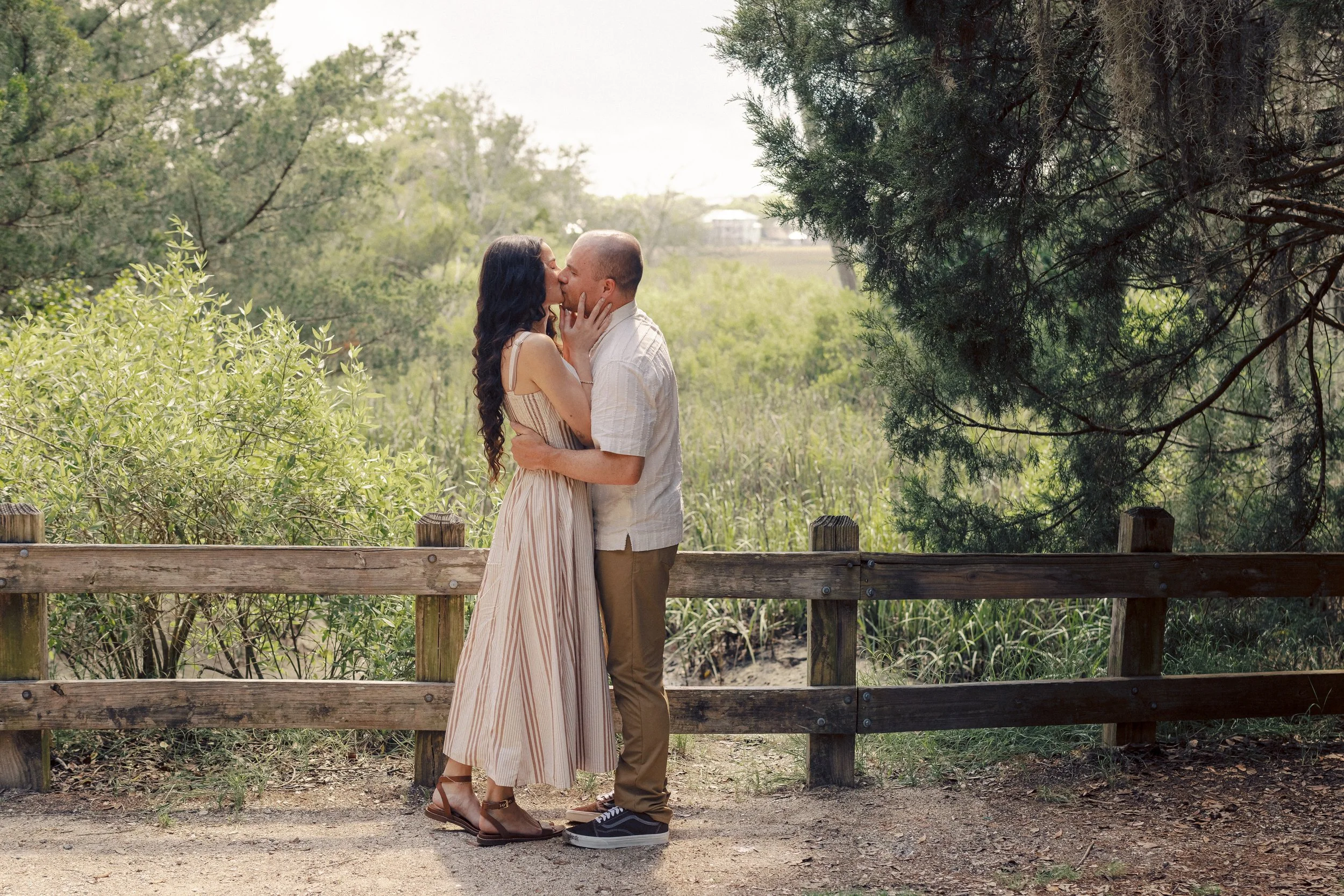 A couple sharing a kiss outdoors on a wooden pathway surrounded by green trees and bushes.
