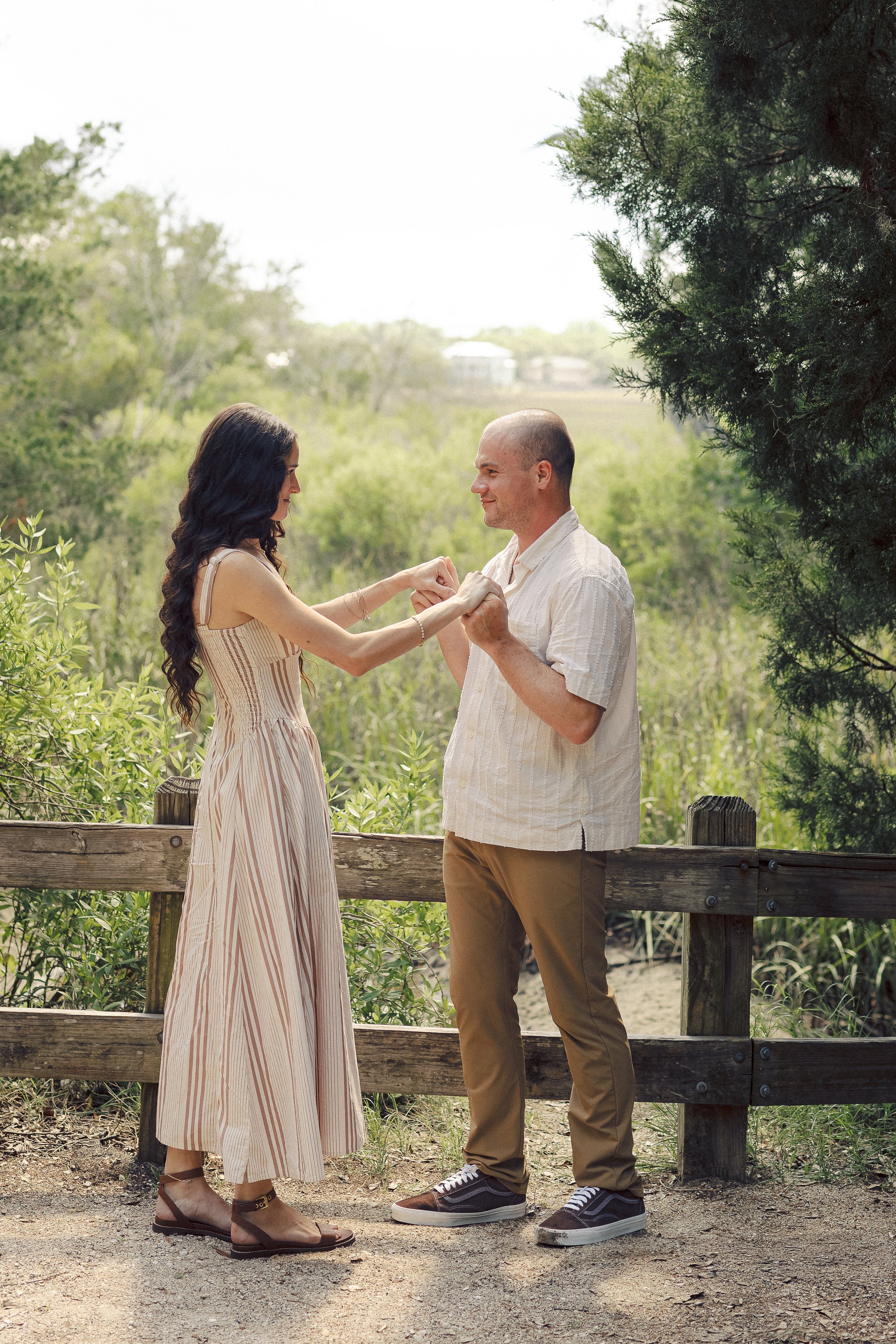 A couple standing outdoors near a wooden fence, holding hands and smiling at each other. The woman has long dark hair and is wearing a beige striped dress with ankle-strap sandals. The man has a shaved head and is dressed in a light-colored button-up