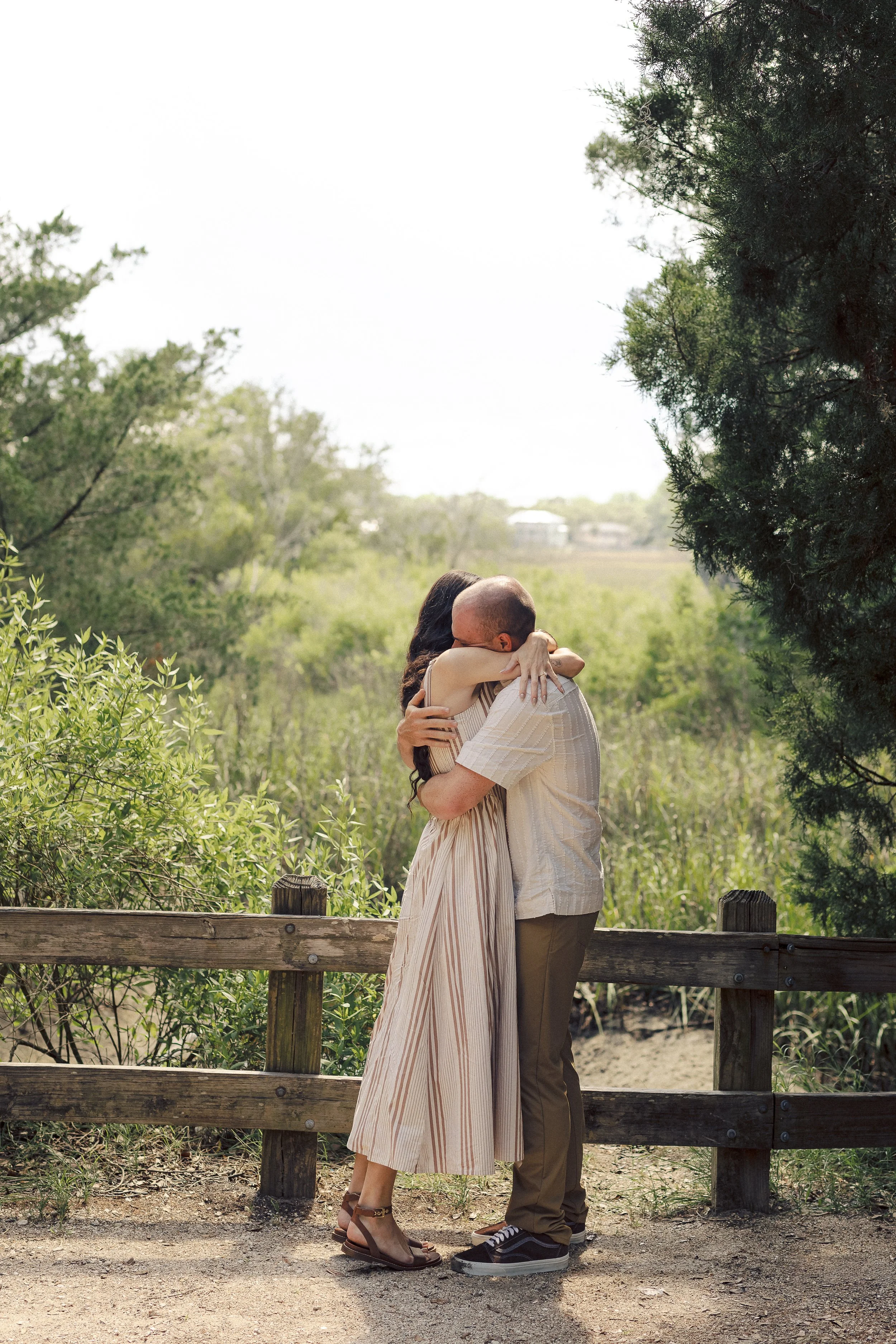 A couple hugging on a wooden path in a green park or nature reserve, with trees and shrubs surrounding them and a bright clear sky overhead.
