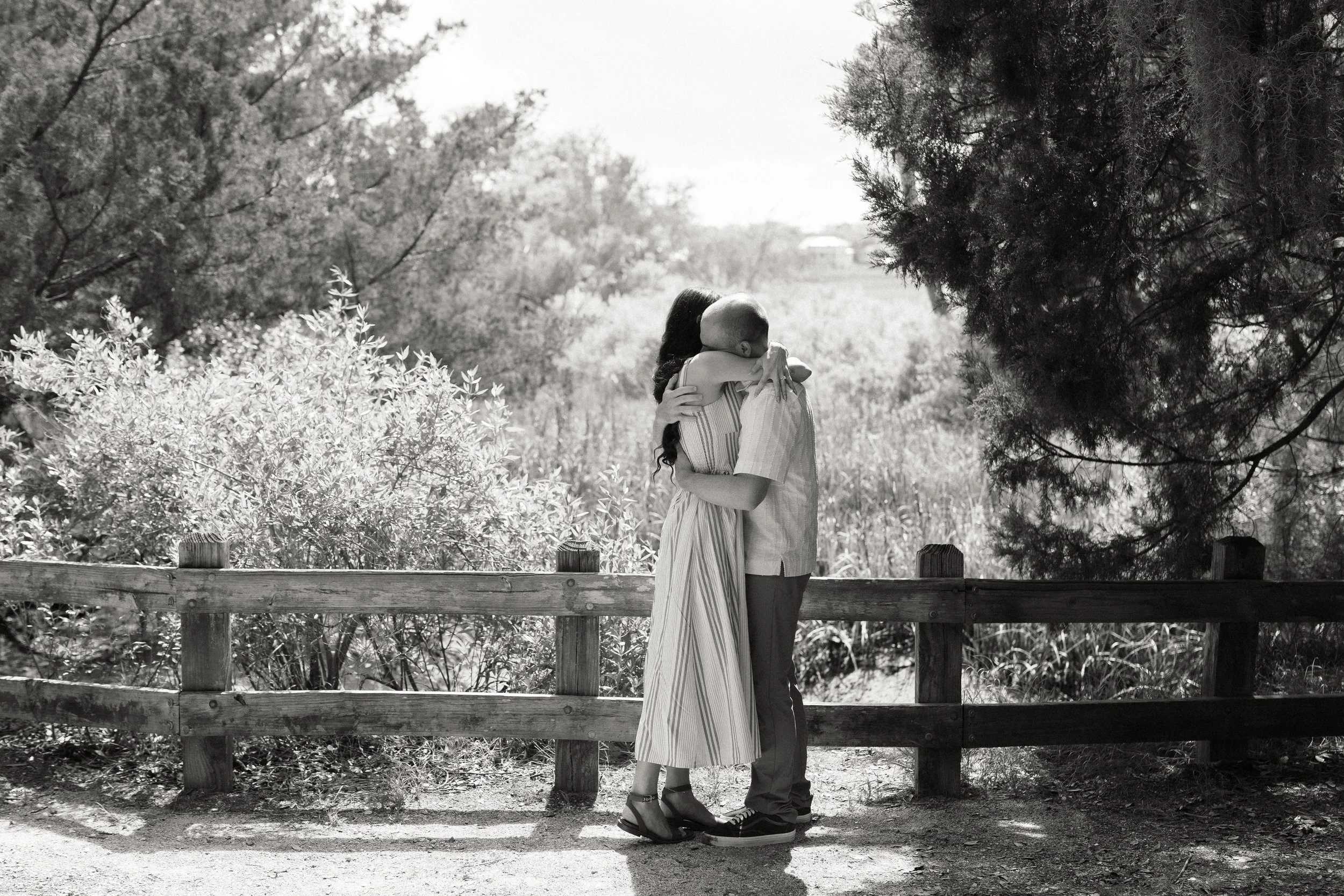 A black and white photo of a couple hugging on a wooden bridge in a natural outdoor setting with trees and bushes.