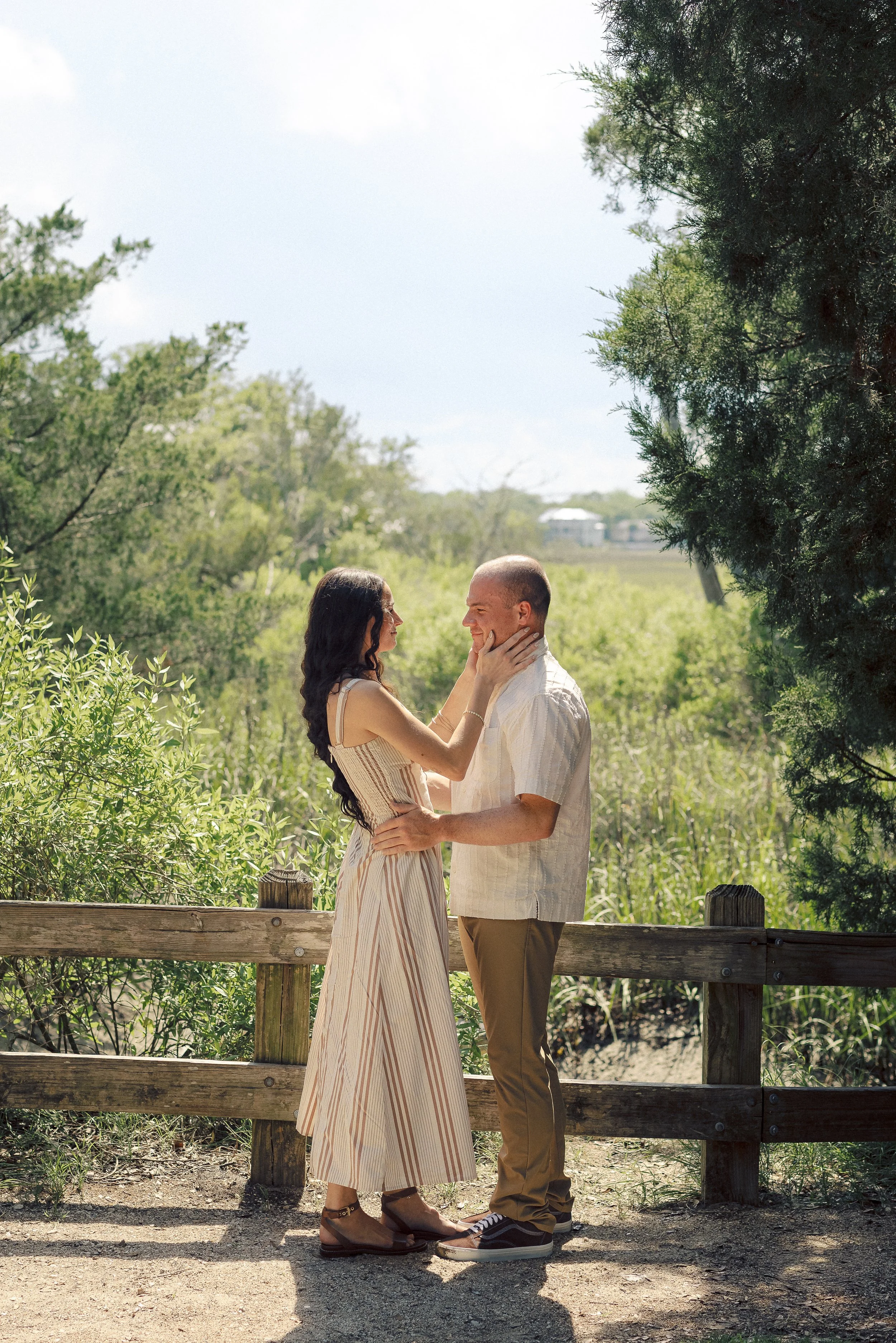 A couple standing close together outdoors, gazing into each other's eyes, with trees and greenery in the background.
