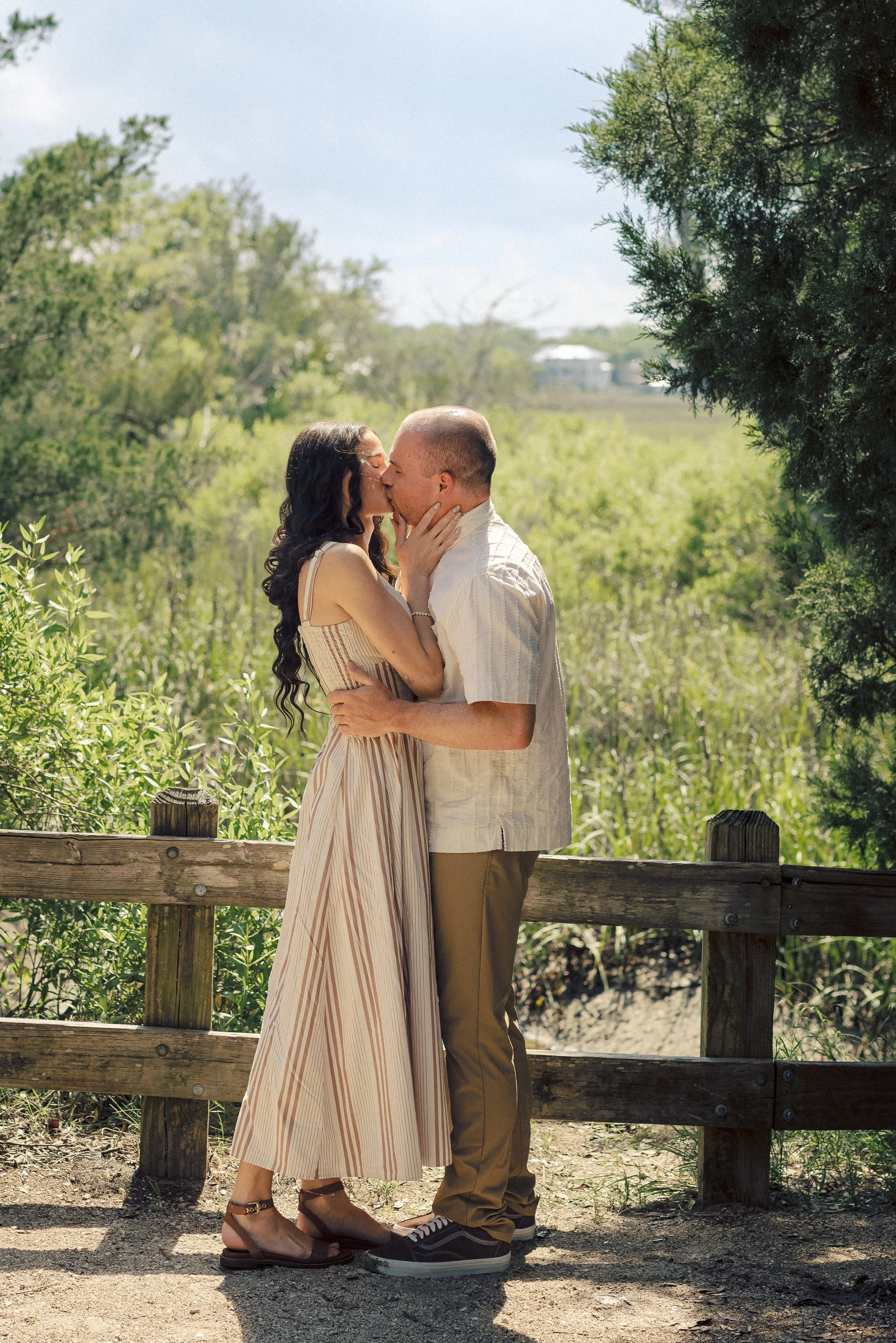 A couple kissing outdoors on a sunny day, standing on a gravel path by a wooden fence surrounded by greenery and trees.