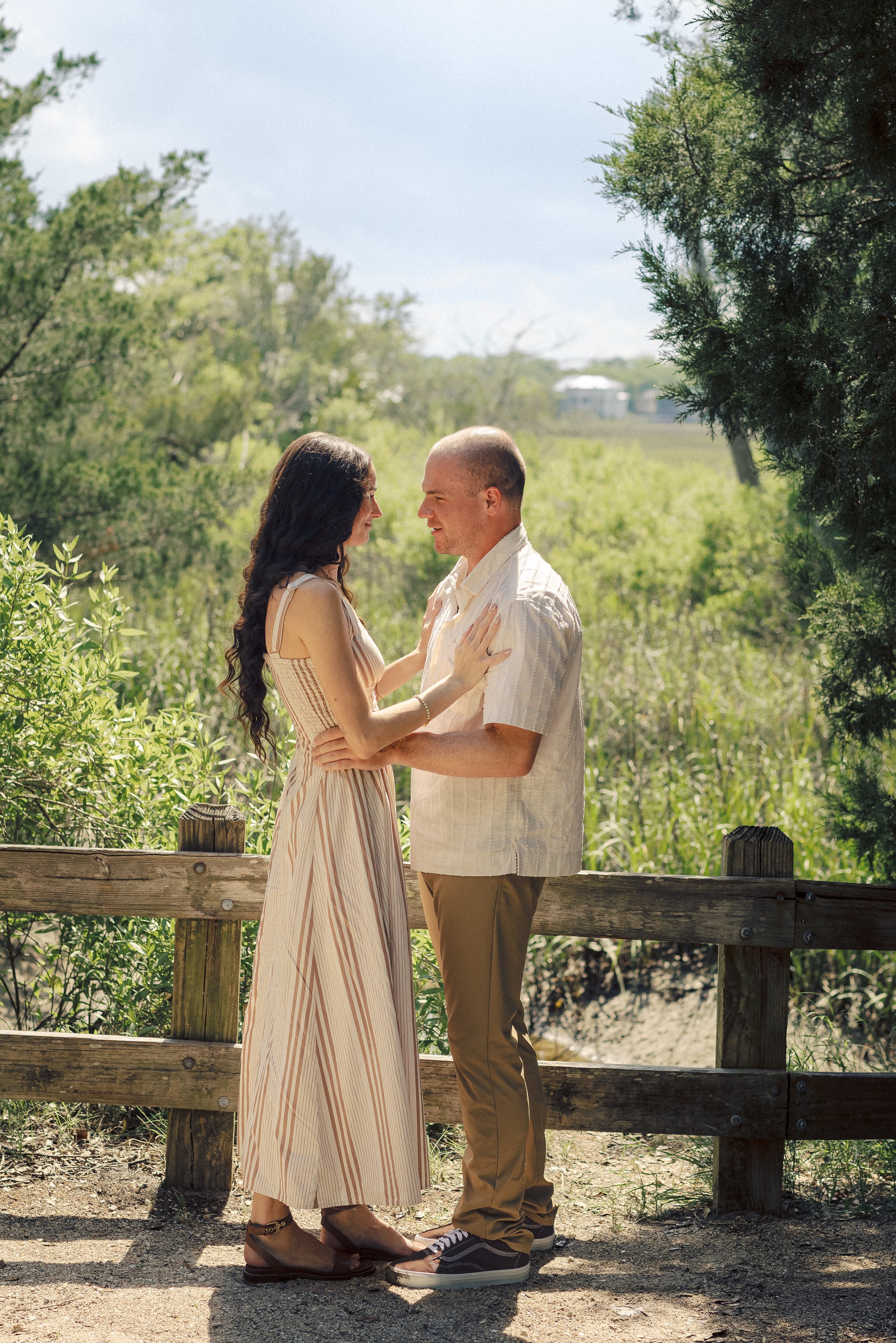 A young couple stands close together facing each other on a sunny day, with the woman touching the man's chest. They are outdoors near a wooden railing, surrounded by greenery and trees, with a building in the background.