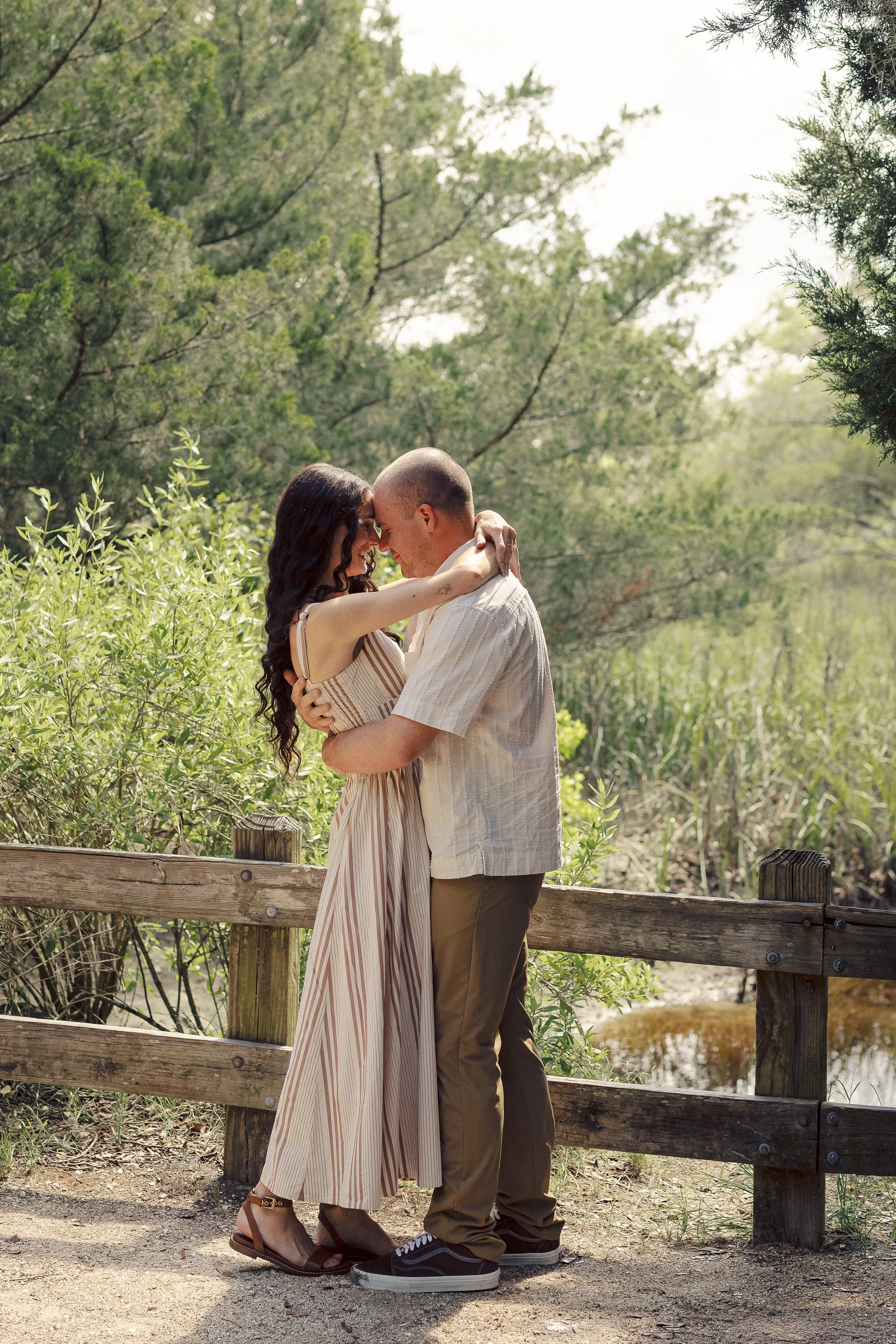 A couple standing closely together with foreheads touching, smiling, and holding each other affectionately outdoors on a sunny day surrounded by green trees and shrubbery.