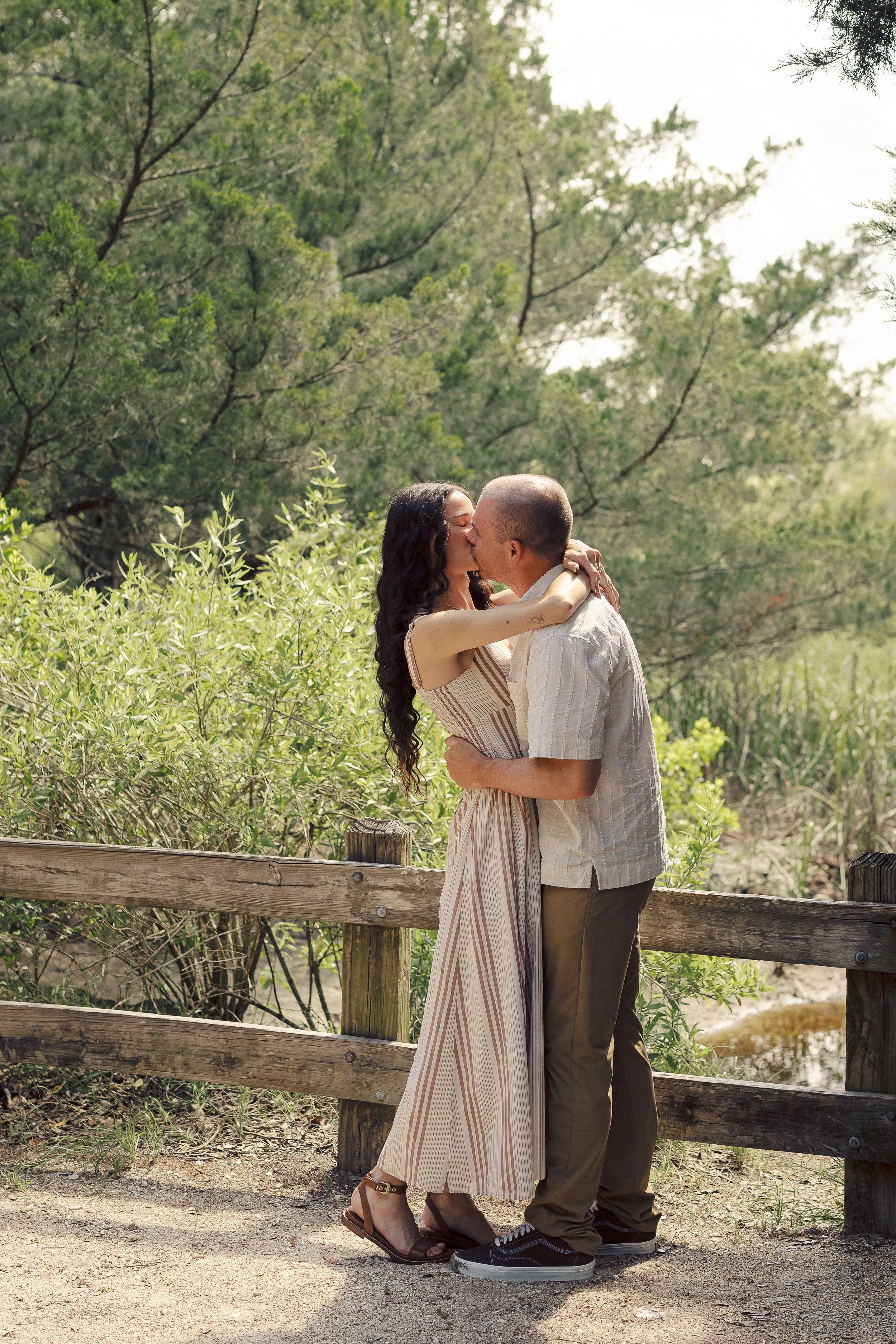 A couple sharing a kiss in a lush outdoor setting with greenery, a wooden fence, and a small stream in the background.
