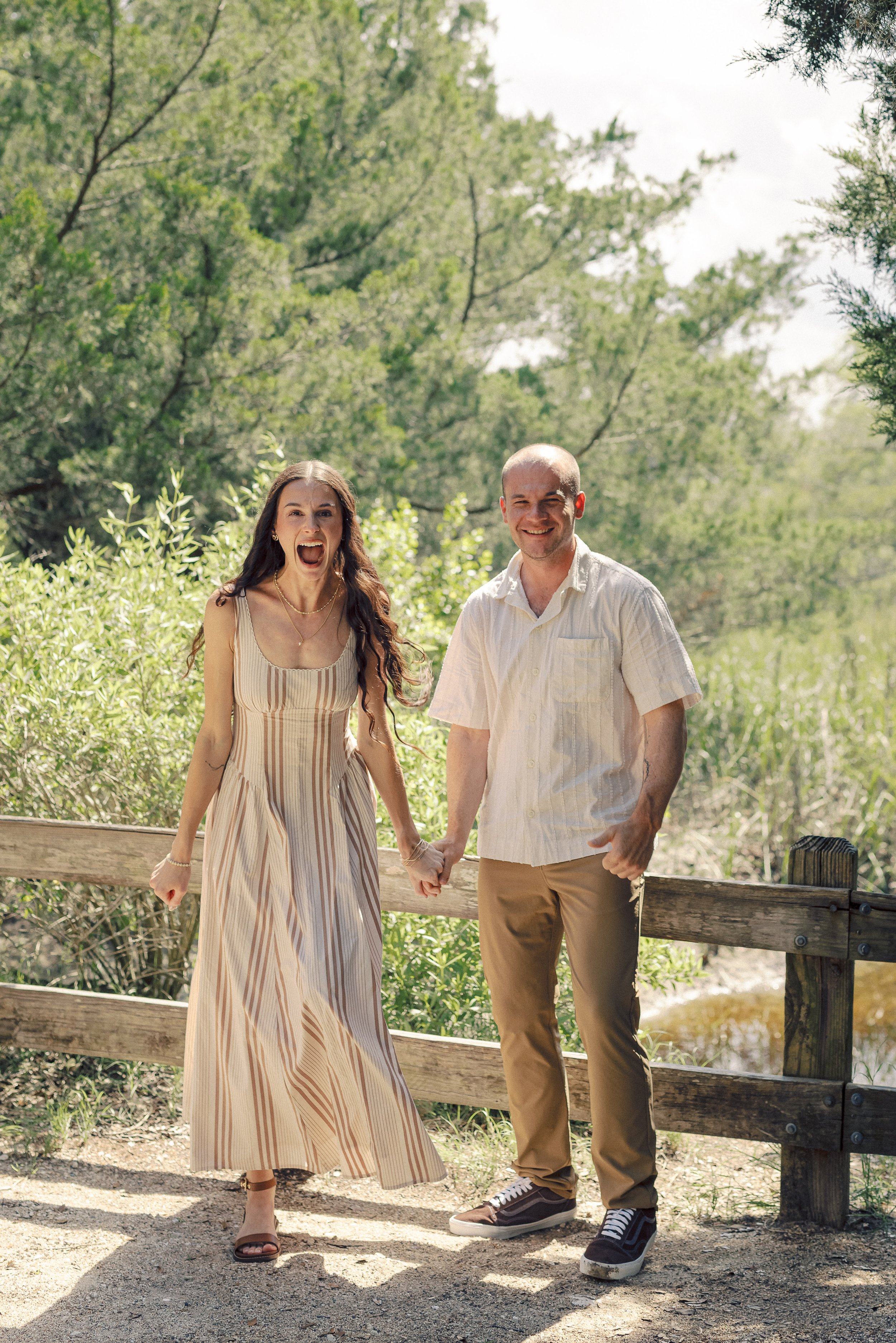 A happy couple holding hands on a wooden bridge in a green outdoor setting, smiling and enjoying the sunny day.