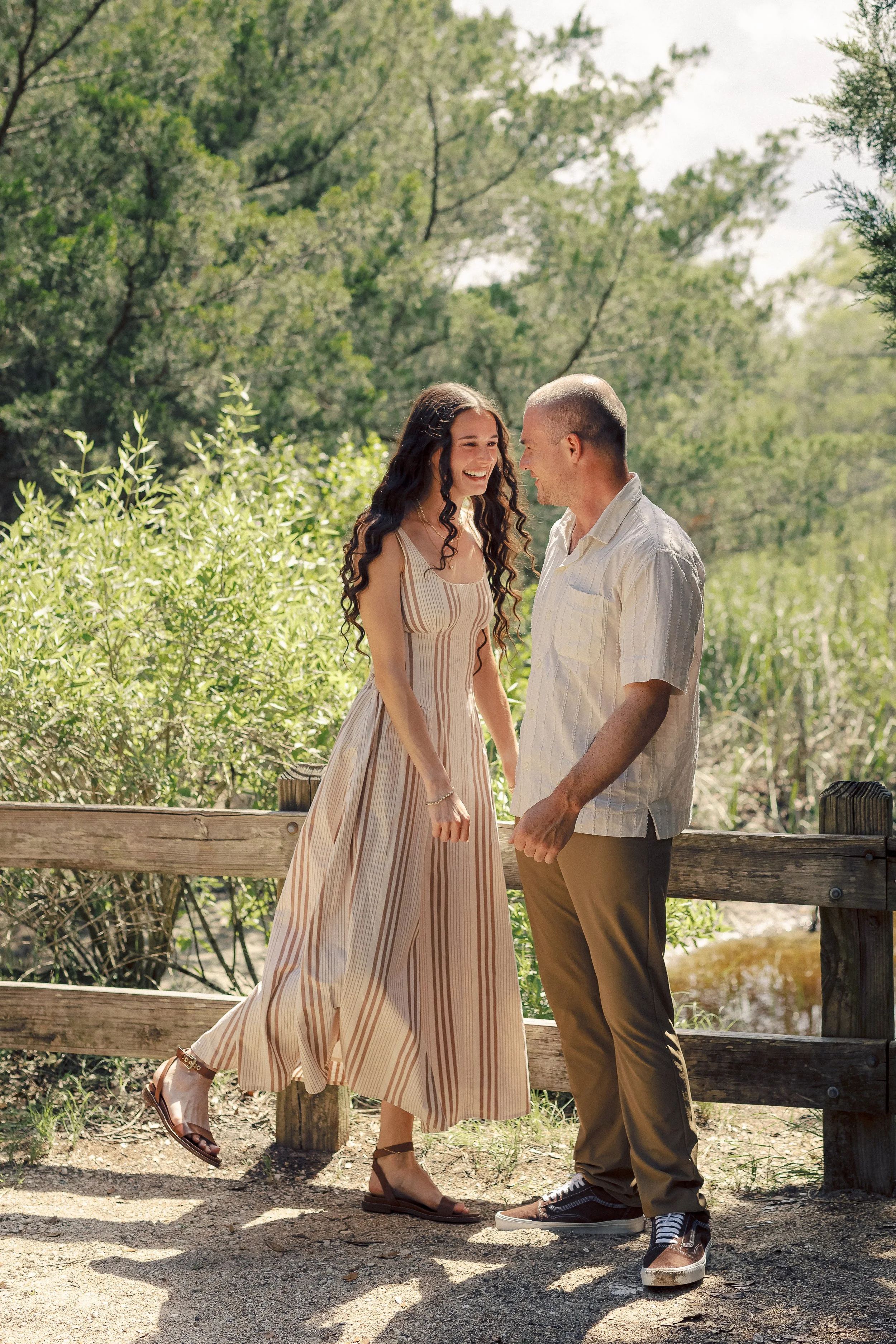 A young woman and a young man are standing on a wooden bridge in a lush, green outdoor setting. The woman is wearing a long, striped beige and white dress with open-toe sandals, and the man is wearing a light-colored short-sleeve shirt, brown pants, 