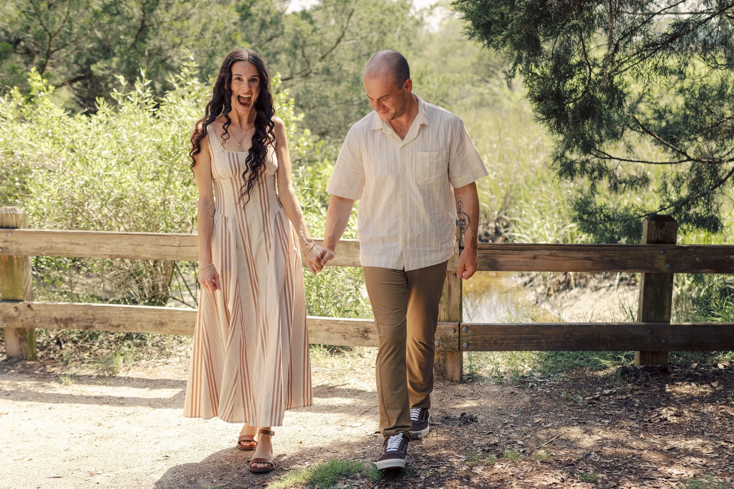 A young woman and a young man walking hand in hand on a dirt path in a wooded area, smiling and enjoying each other's company.