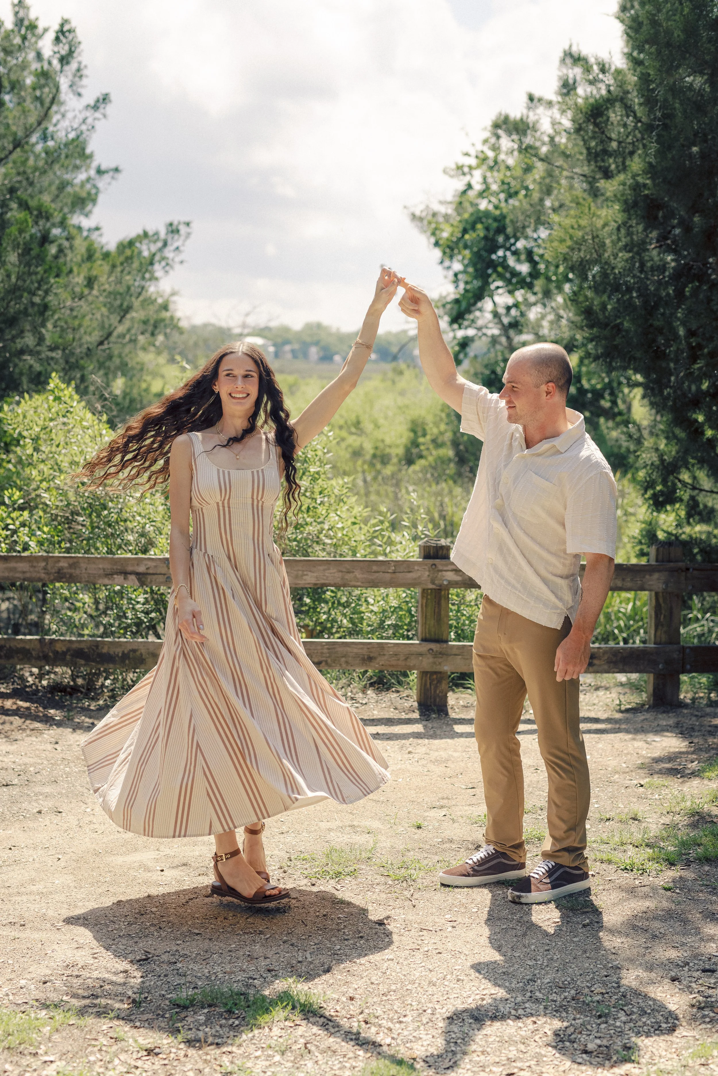 A young woman in a striped, flowing dress and high-heeled sandals dancing outdoors with a young man in casual tan pants and sneakers, holding hands in the air, on a sunny day in a park with trees and a wooden fence in the background.