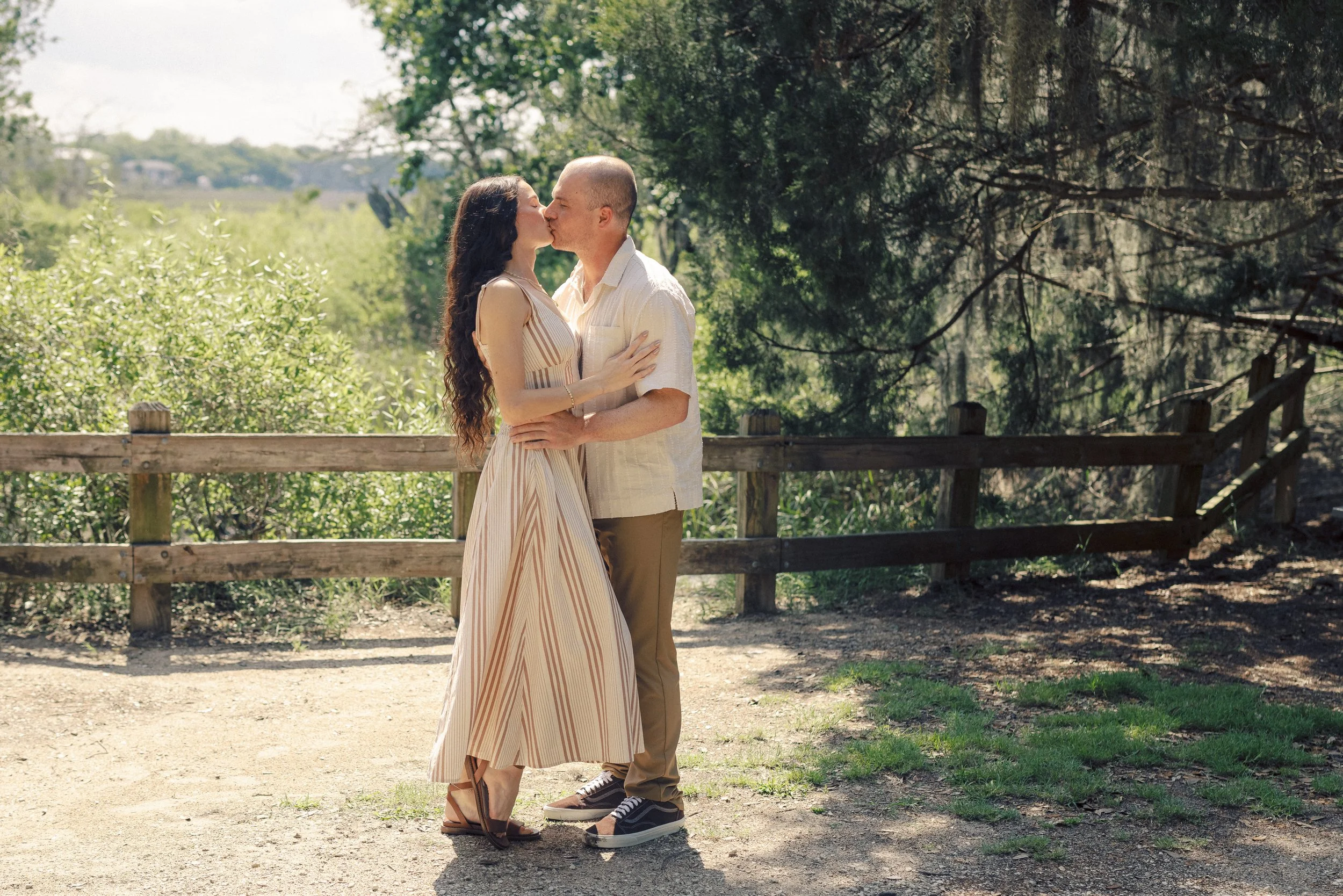 A couple sharing a kiss outdoors near a wooden fence, surrounded by green trees and bushes, under a partly sunny sky.