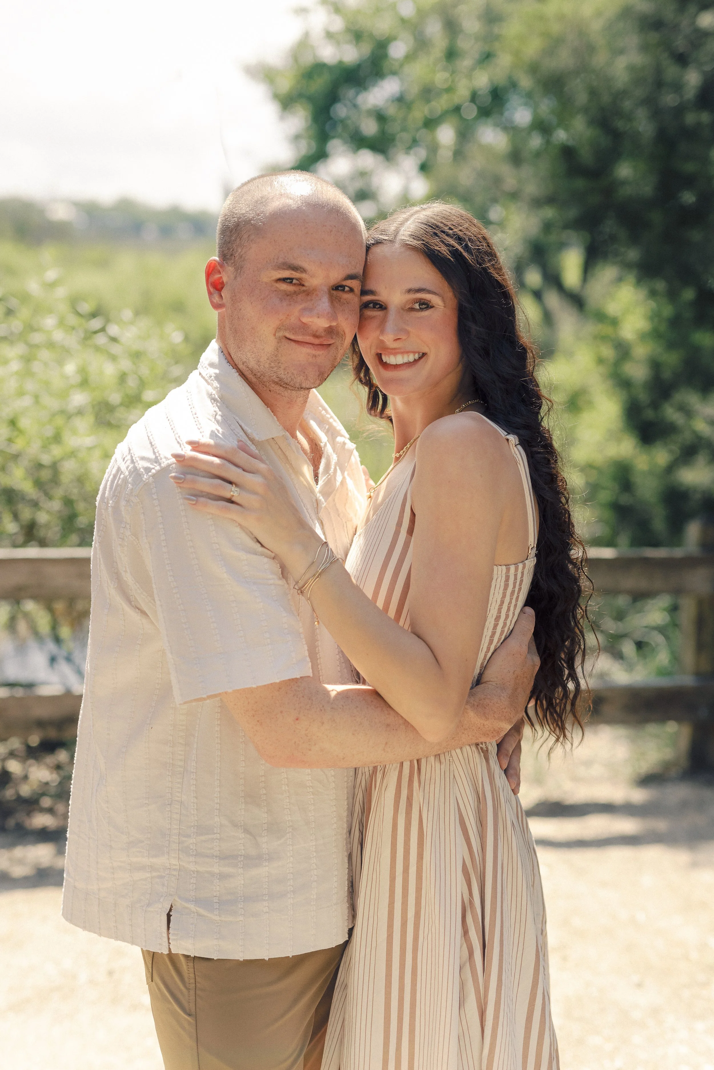 A couple standing outdoors, embracing each other and smiling at the camera. The setting is sunny with greenery in the background.