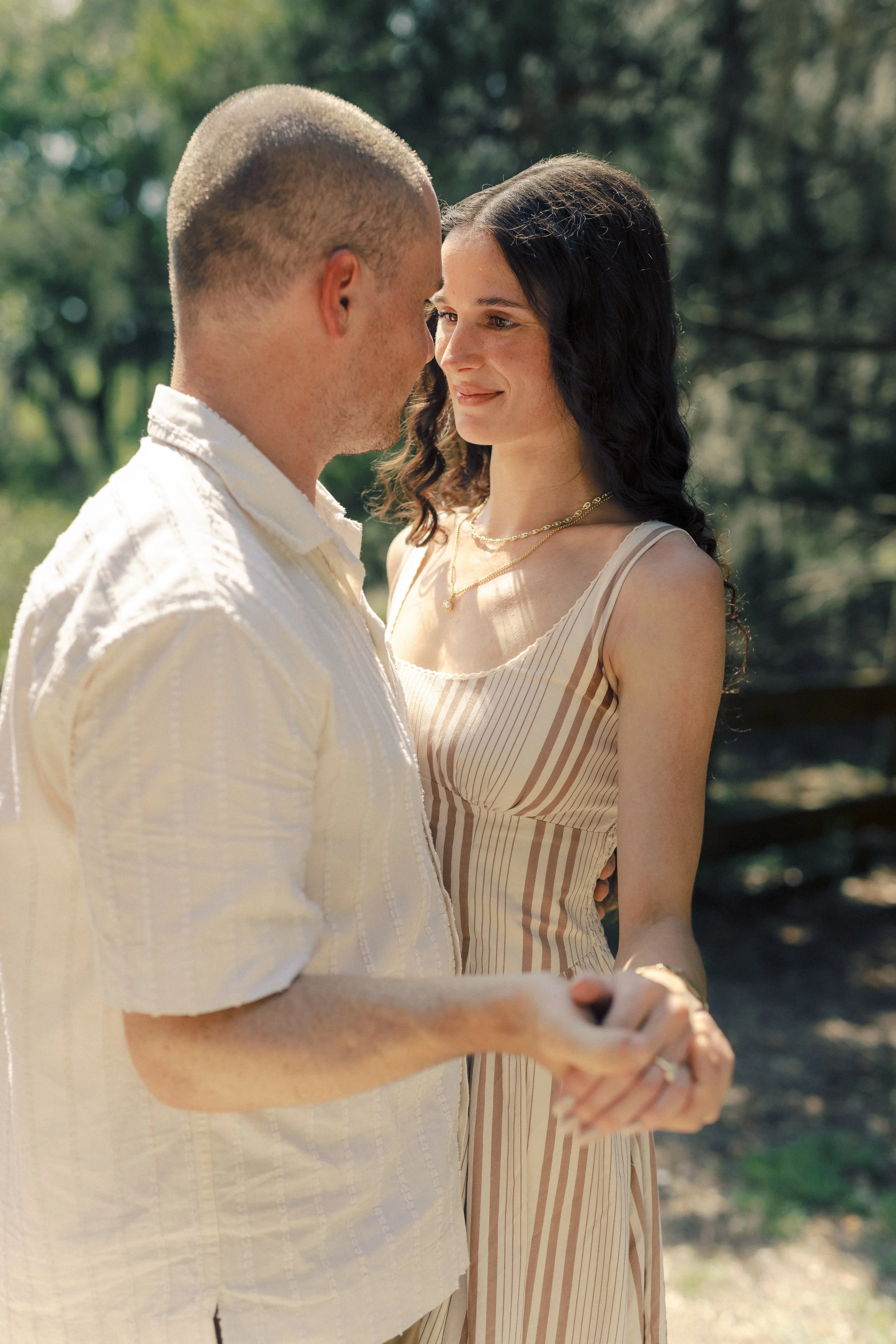 A young couple holding hands outdoors with trees in the background, facing each other and smiling gently.