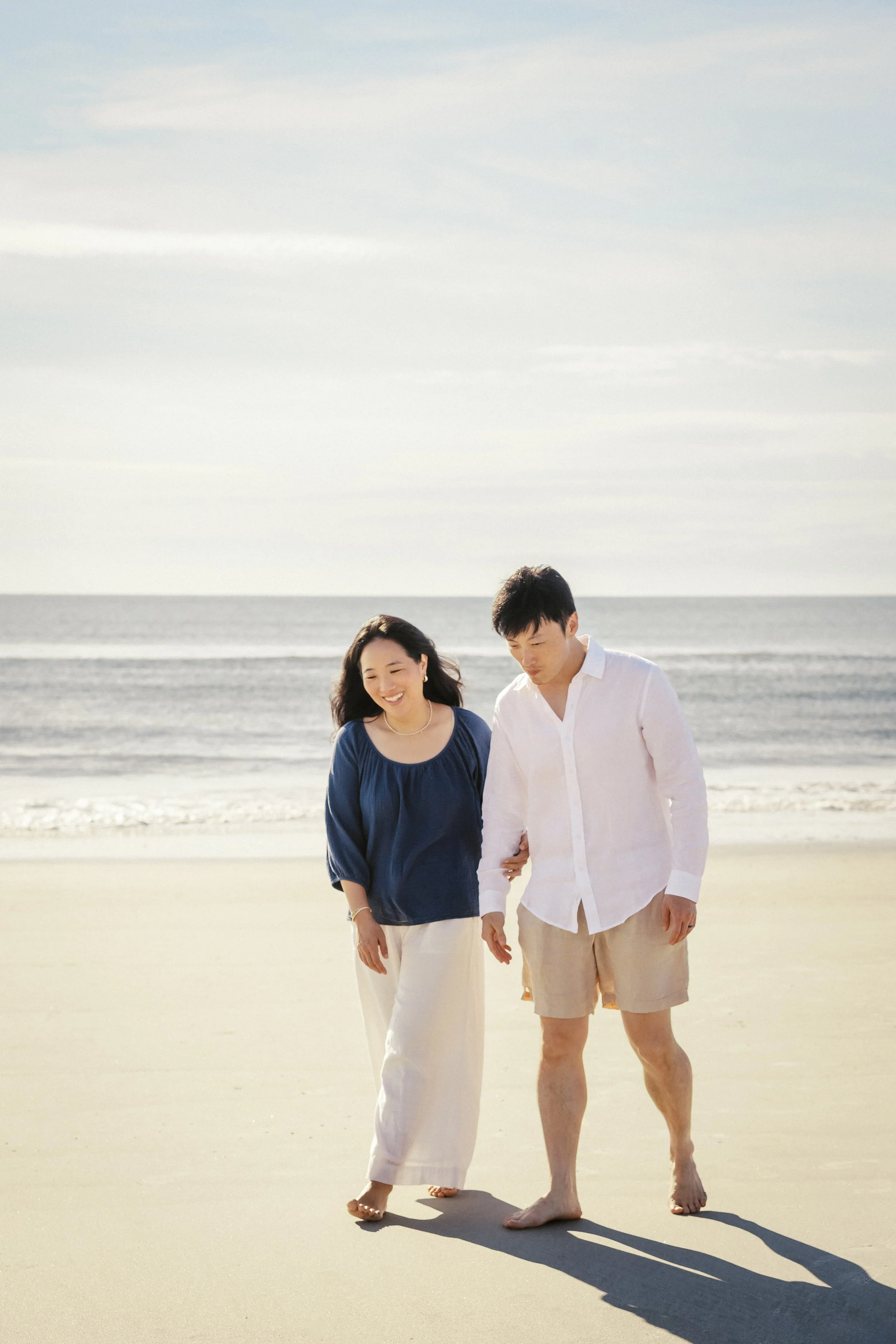 A smiling woman and a man holding hands walking along the beach near the ocean, with a cloudy sky overhead.Photographer Amelia Island, Jacksonville, St Augustine, St Simons Island, Savannah photographer