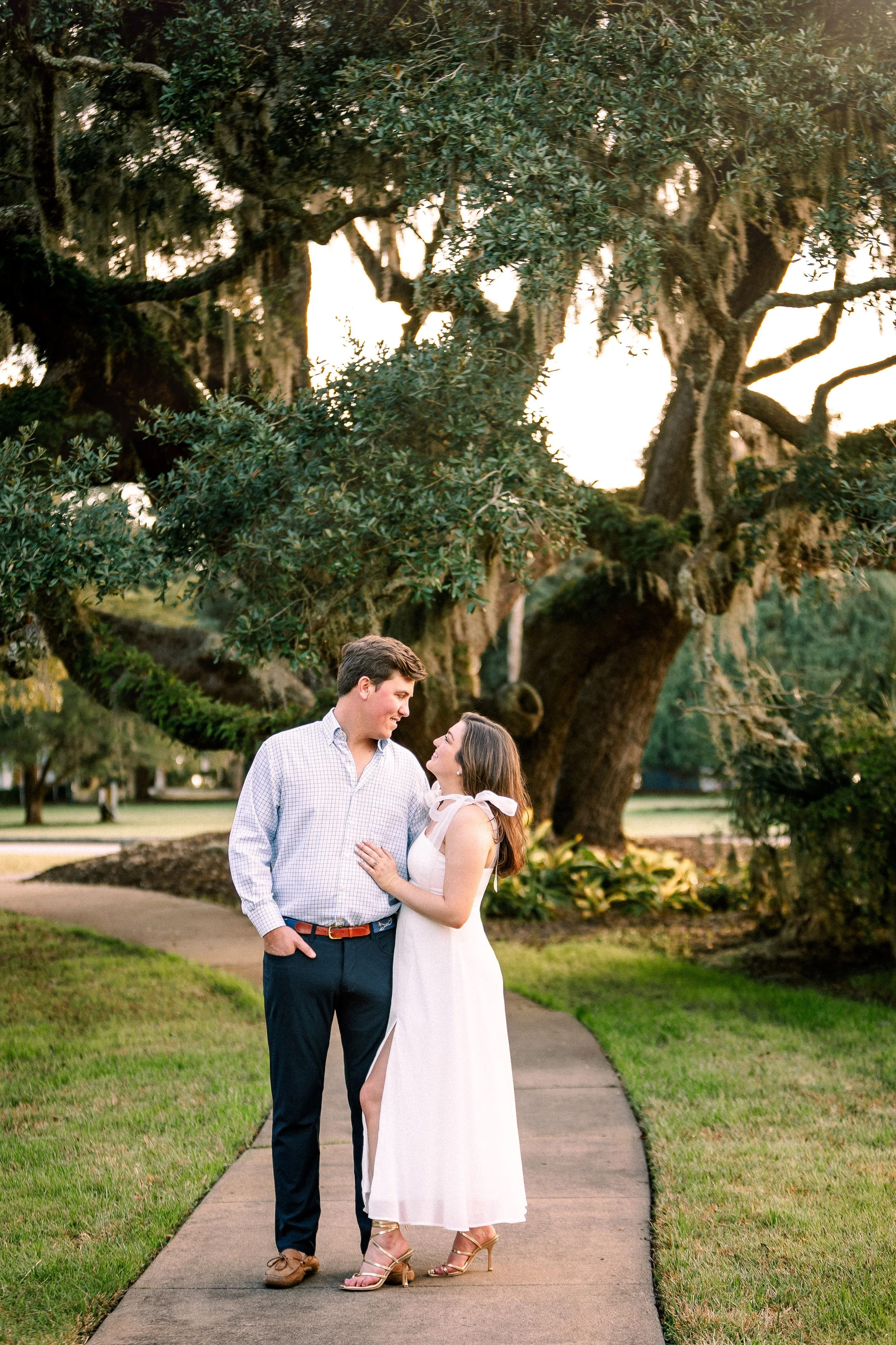A man and woman standing close together, looking into each other's eyes, on a sidewalk in a park with large trees and green grass in the background during sunset.