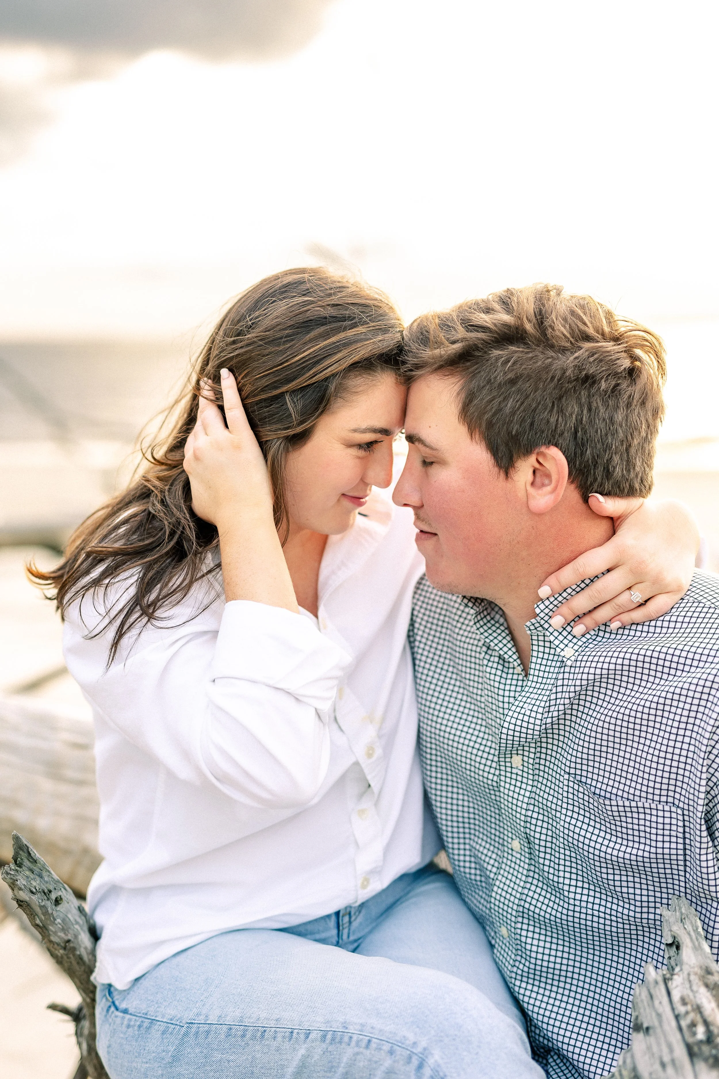 A couple sitting close together outdoors on a beach, with their foreheads touching and eyes closed, sharing an intimate moment.