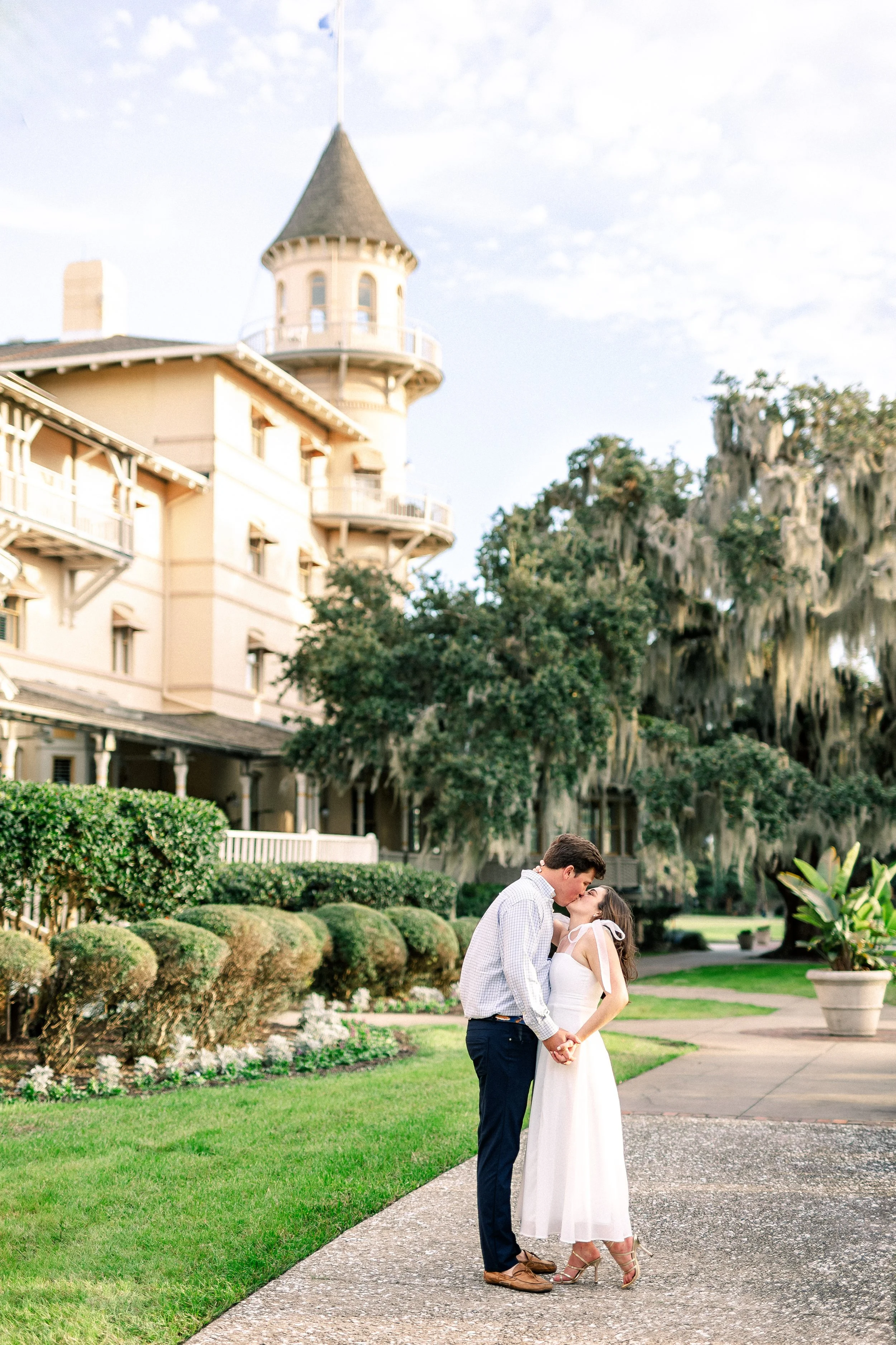 A couple sharing a kiss outdoors on a walking path, with a large house and greenery in the background.