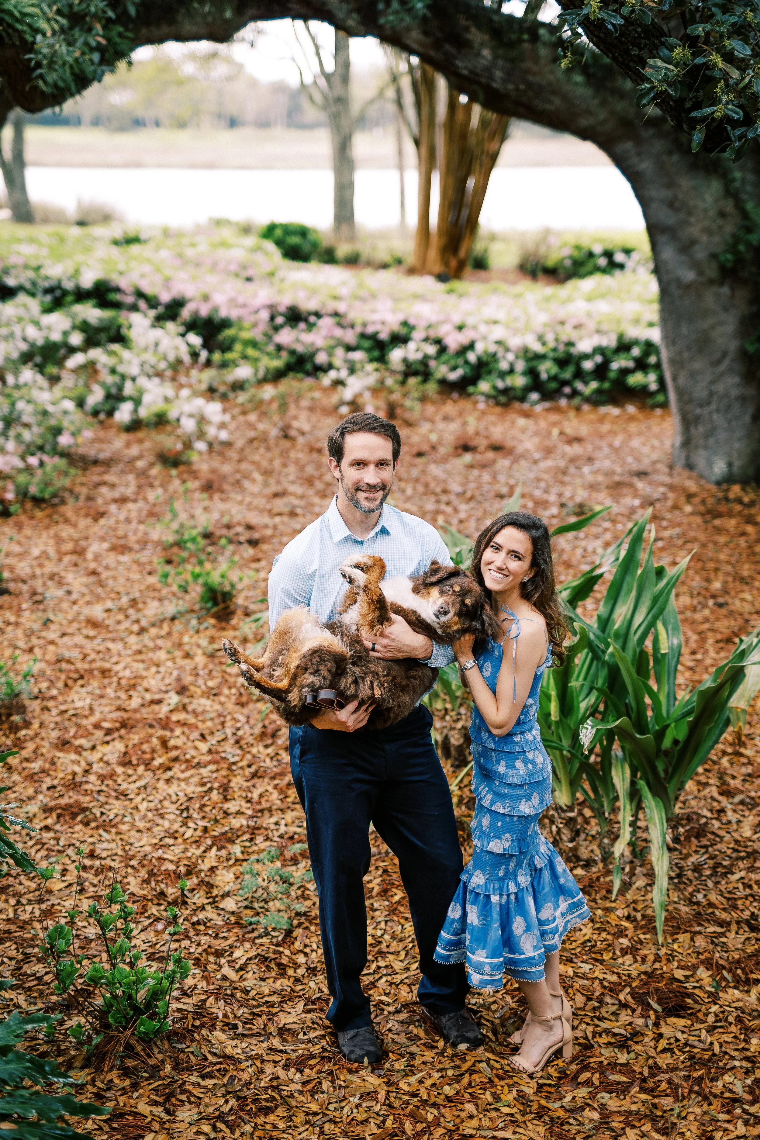 A man and woman standing together outdoors on a leaf-covered ground, holding a large group of dogs, smiling at the camera, with trees and flowering bushes in the background.