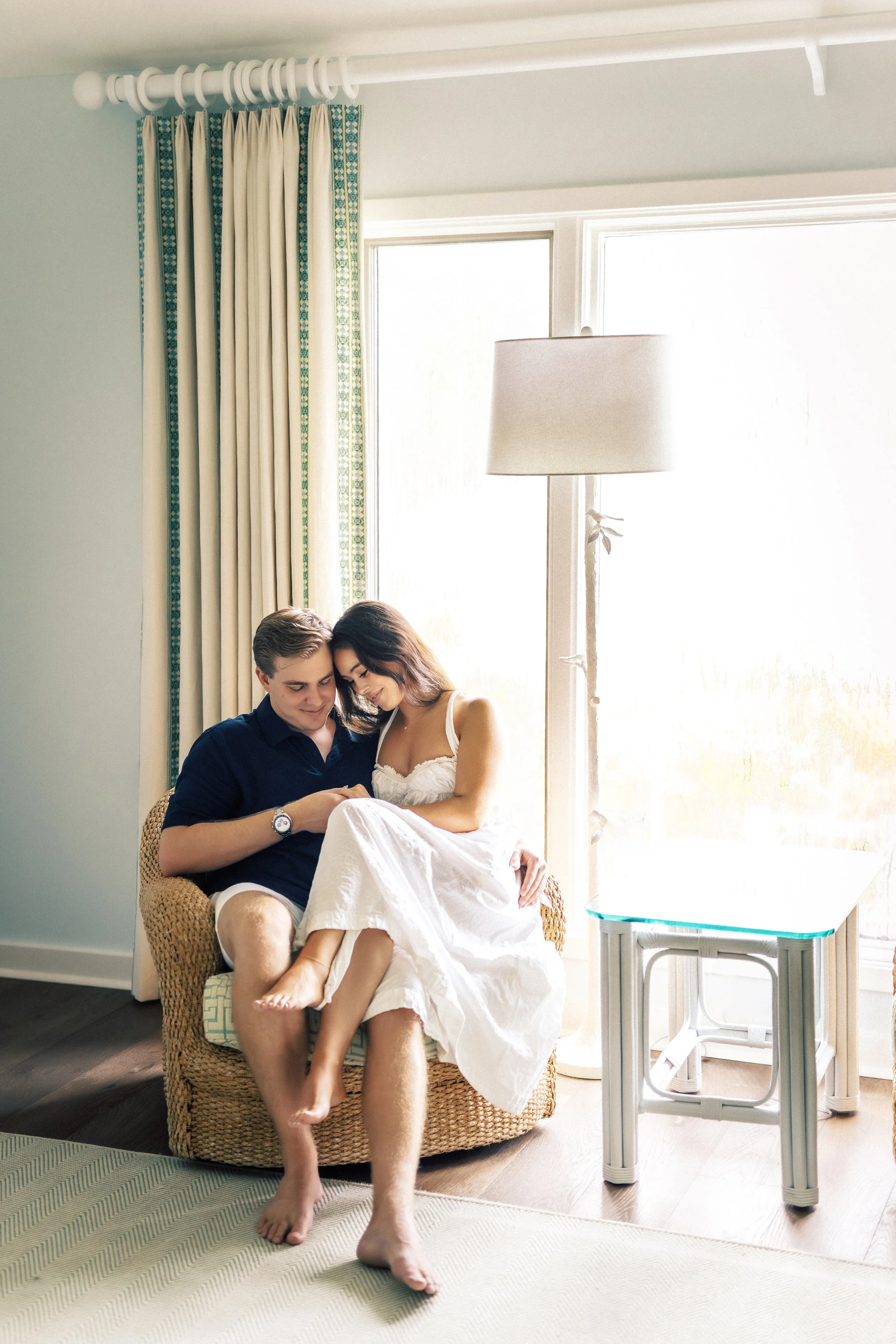 A young couple sitting on a wicker chair near a large window in a bright room, looking at a phone together.