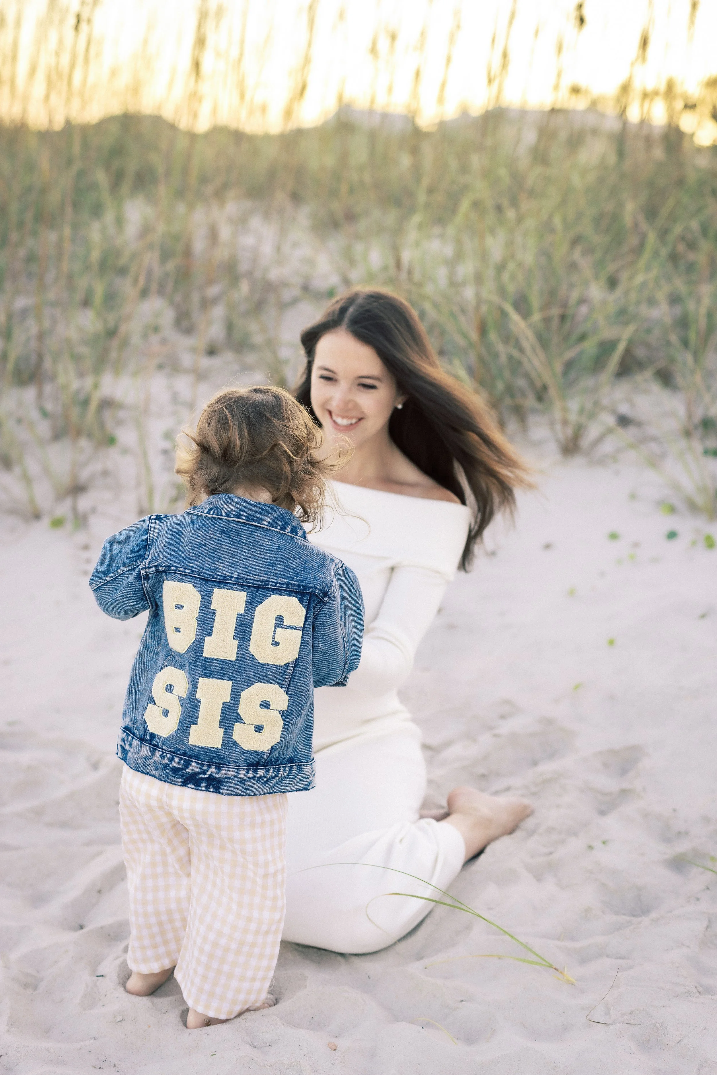 A woman sitting on a sandy beach with a child in front of her, who is facing away from the camera. Photographer Amelia Island, Jacksonville, St Augustine, St Simons Island, Savannah photographer