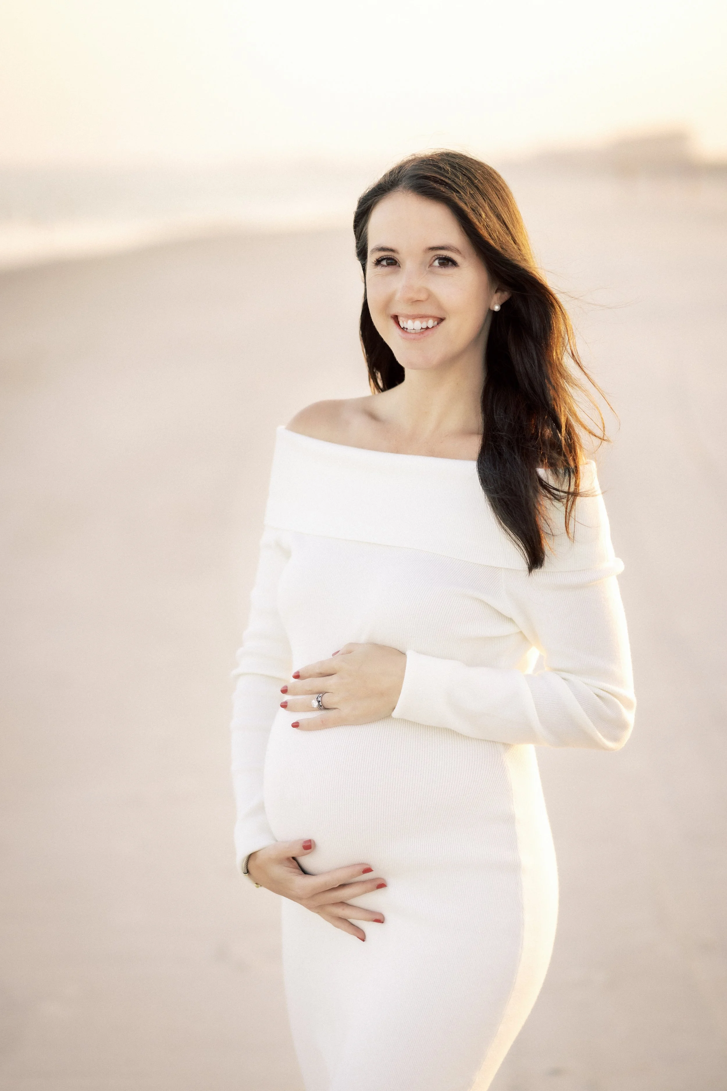 A pregnant woman in a white off-the-shoulder dress standing on a beach, smiling and holding her belly with her left hand.Photographer Amelia Island, Jacksonville, St Augustine, St Simons Island, Savannah photographer