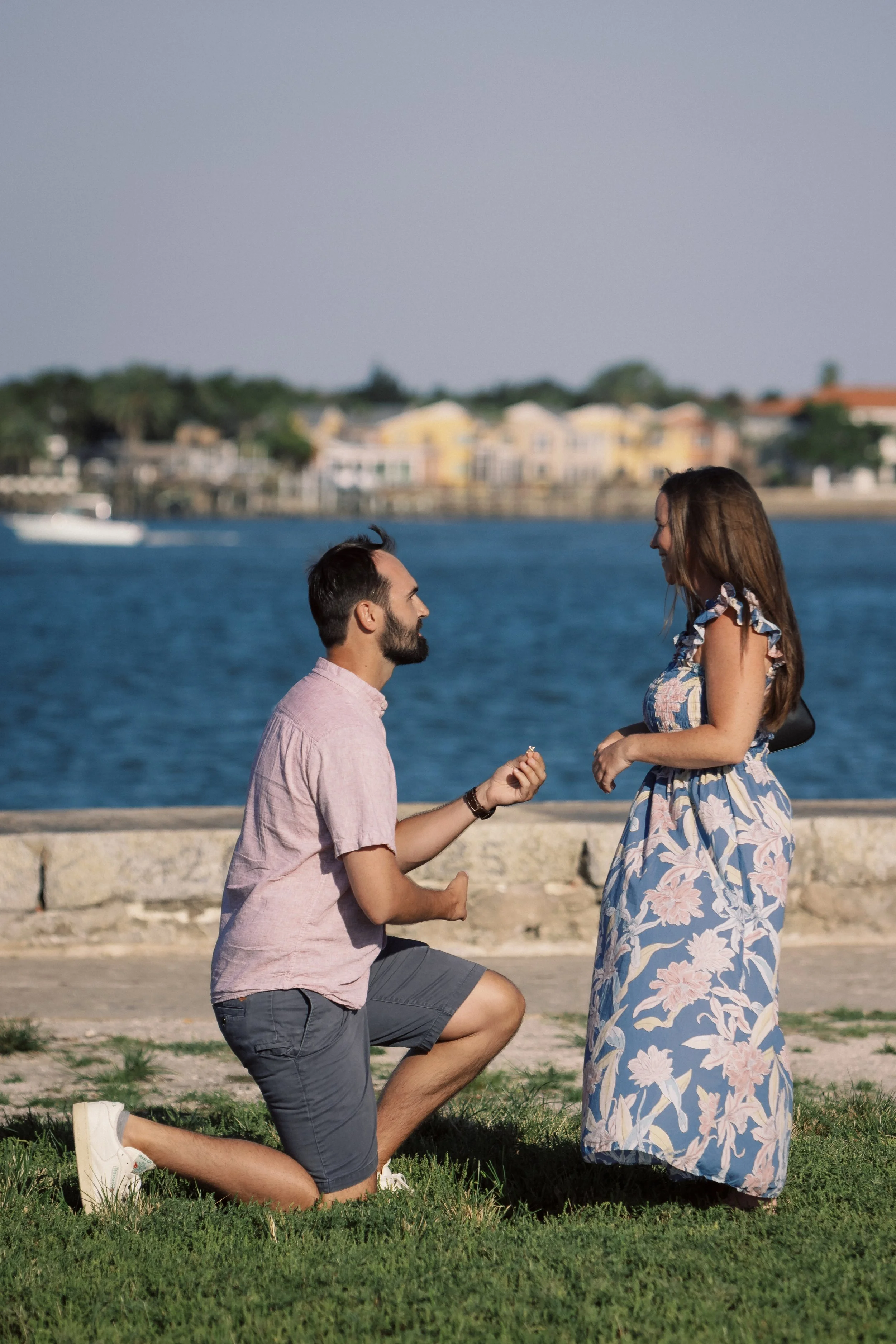 A man kneeling on one knee proposing marriage to a woman by a body of water, with houses and trees in the background.
