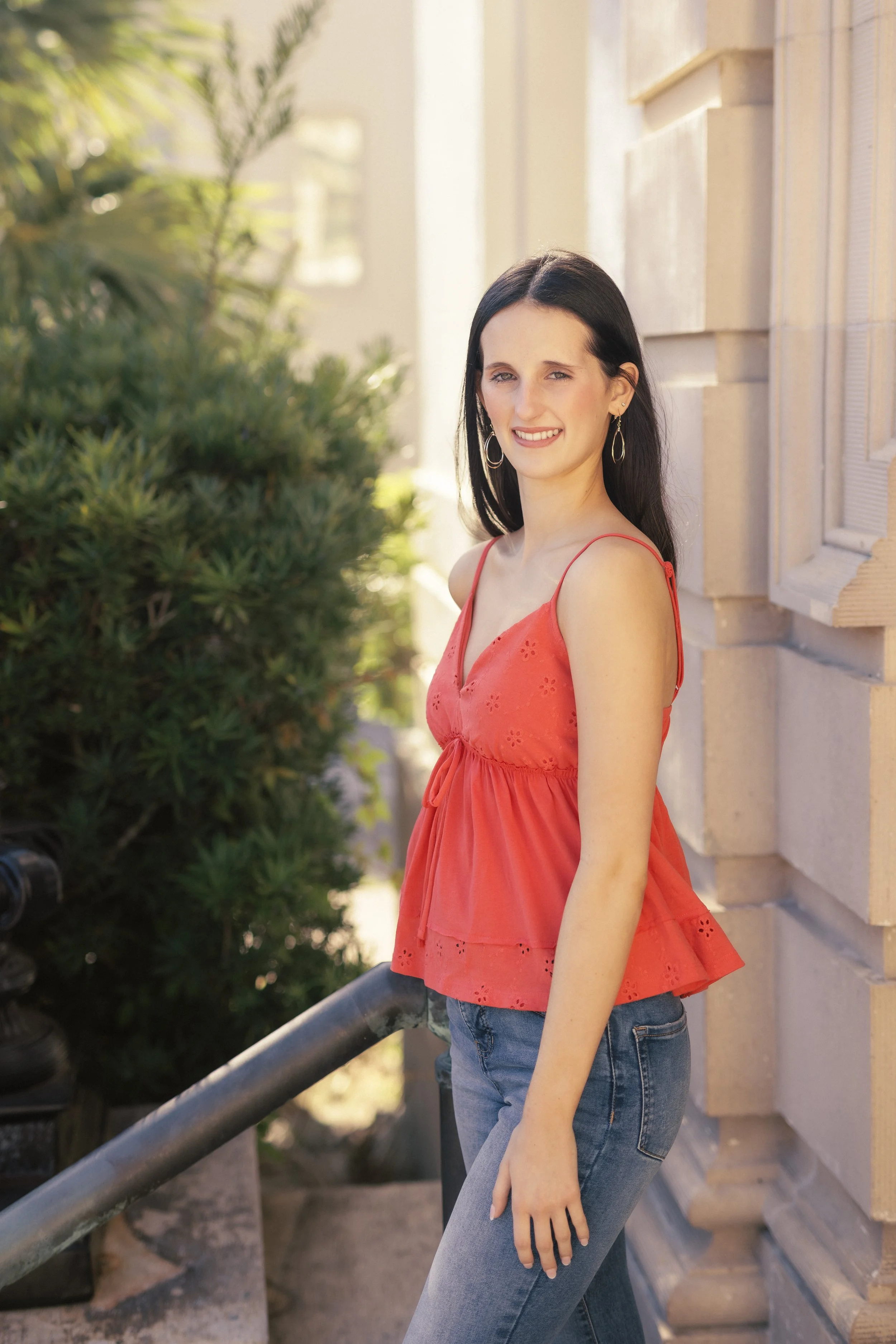 A woman with long dark hair wearing a red sleeveless top and blue jeans standing outside next to a stone building, with greenery in the background.
