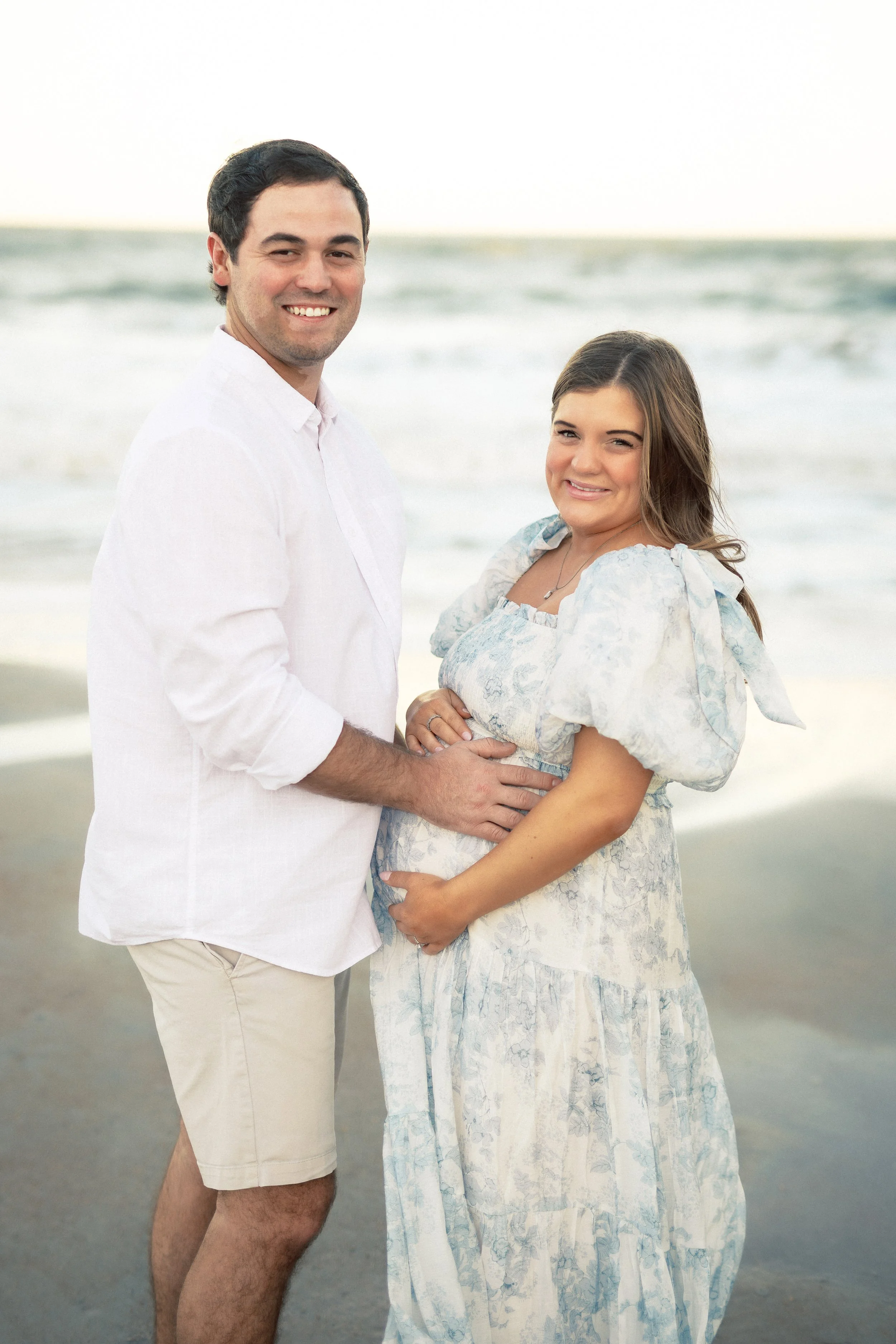 A pregnant woman and a man standing on the beach, smiling at the camera with the ocean and sky in the background.