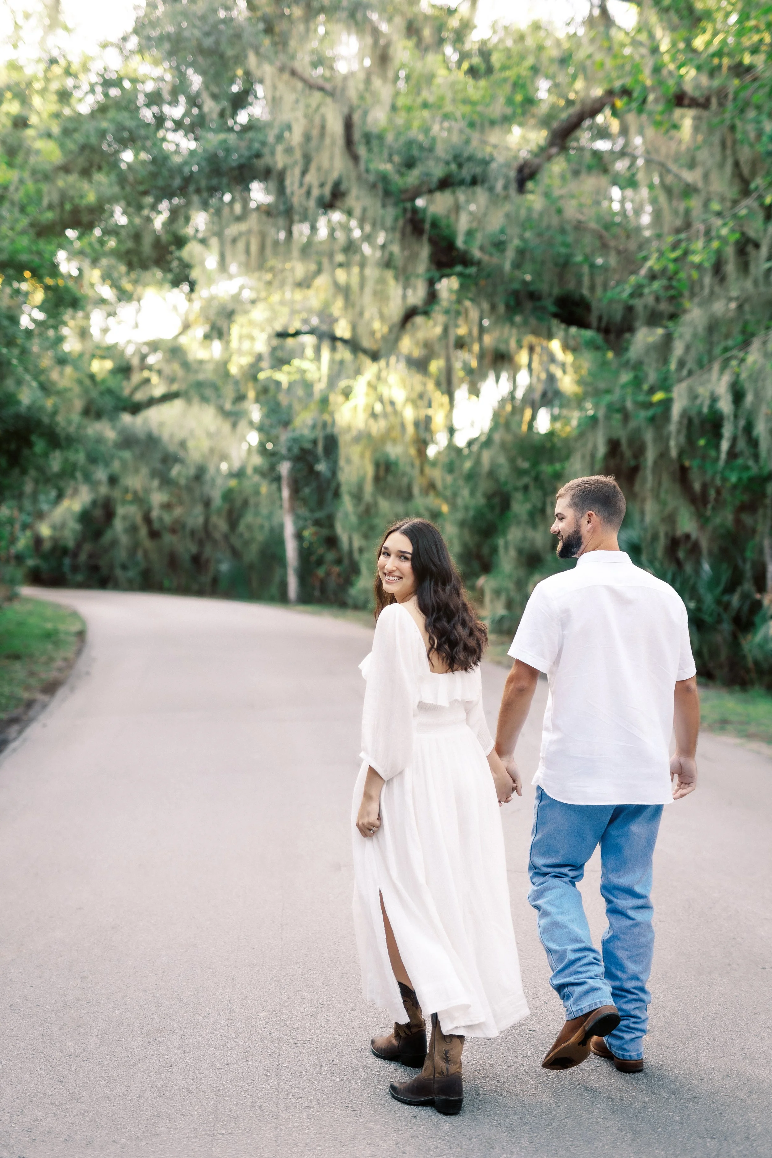 A couple holding hands, walking down a paved path in a lush green park, smiling at each other.