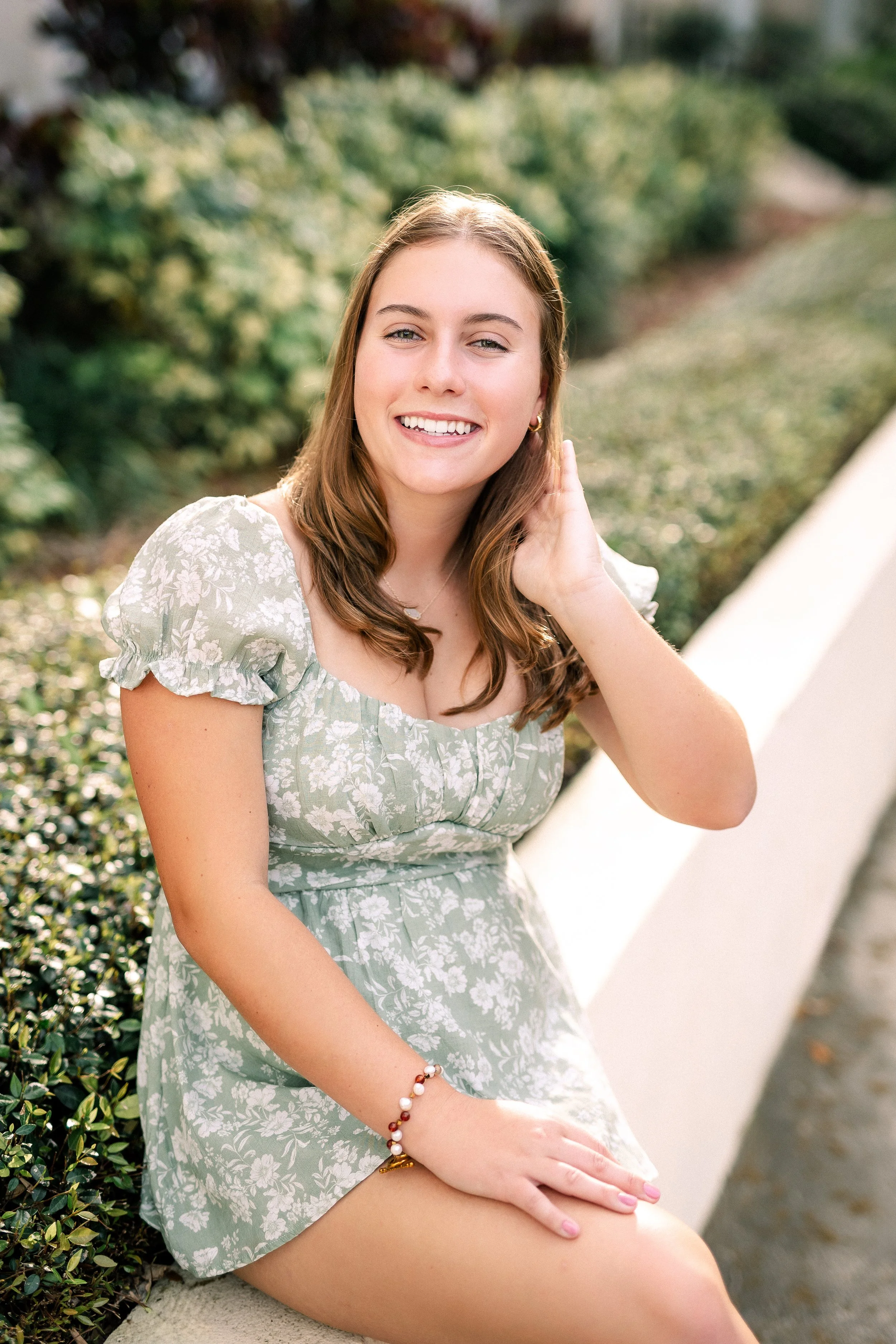 A young woman with light brown hair, smiling, sitting outdoors on a concrete ledge near green foliage, wearing a light green floral dress and a pearl bracelet.