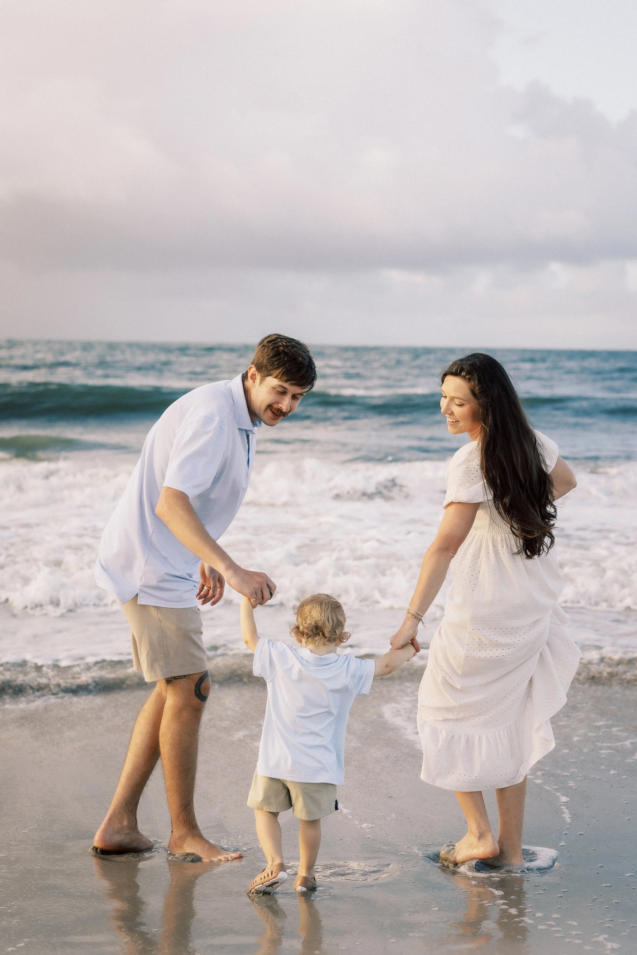 A happy family of four at the beach, with the parents holding their young child's hands as they walk along the shoreline. Photographer Amelia Island, Jacksonville, St Augustine, St Simons Island, Savannah photographer