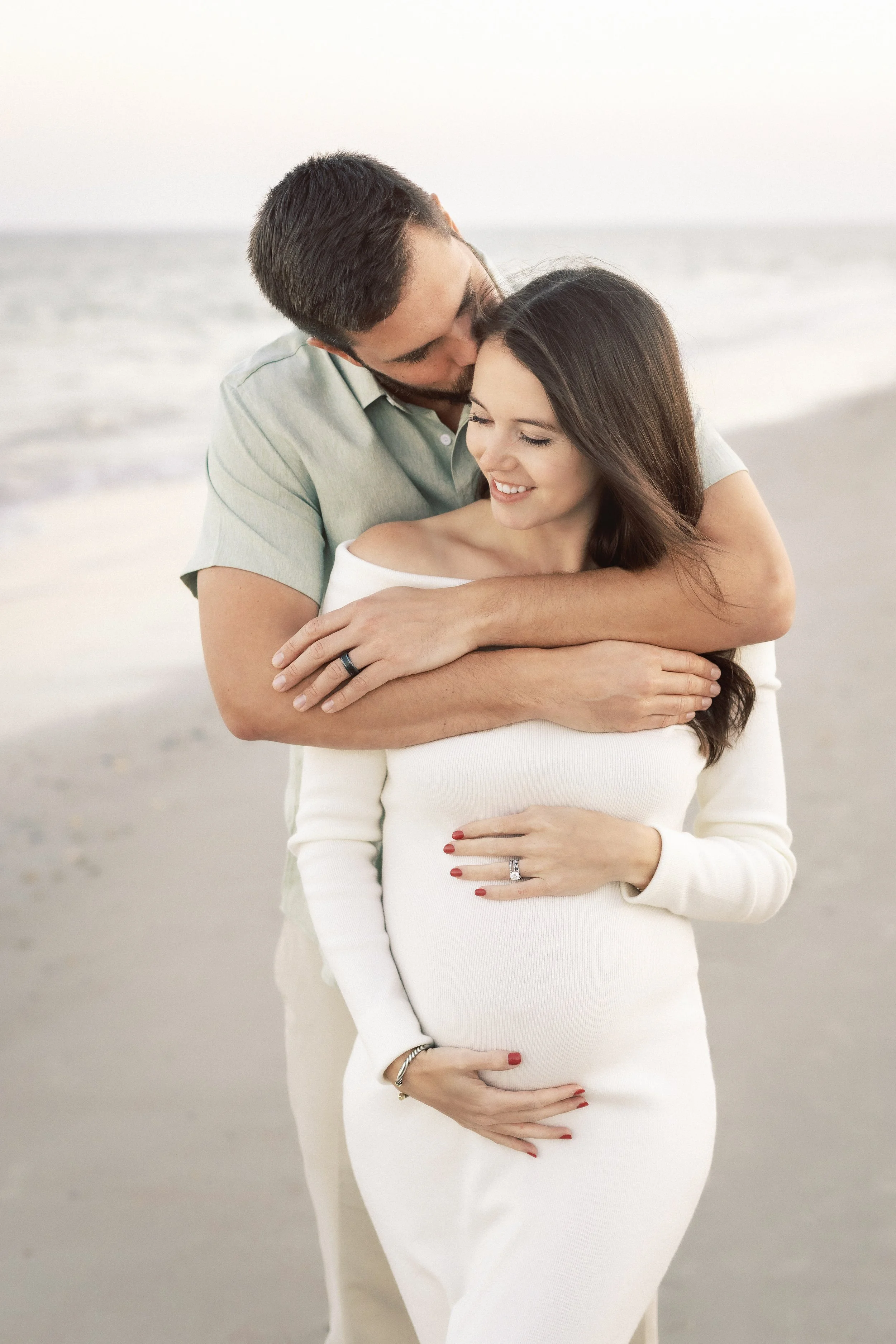 A couple hugging on a beach, with the man kissing the woman's head. The woman is pregnant, wearing a white dress, and showing her baby bump. The man has dark hair and a beard, and the woman has long dark hair. Both are smiling.