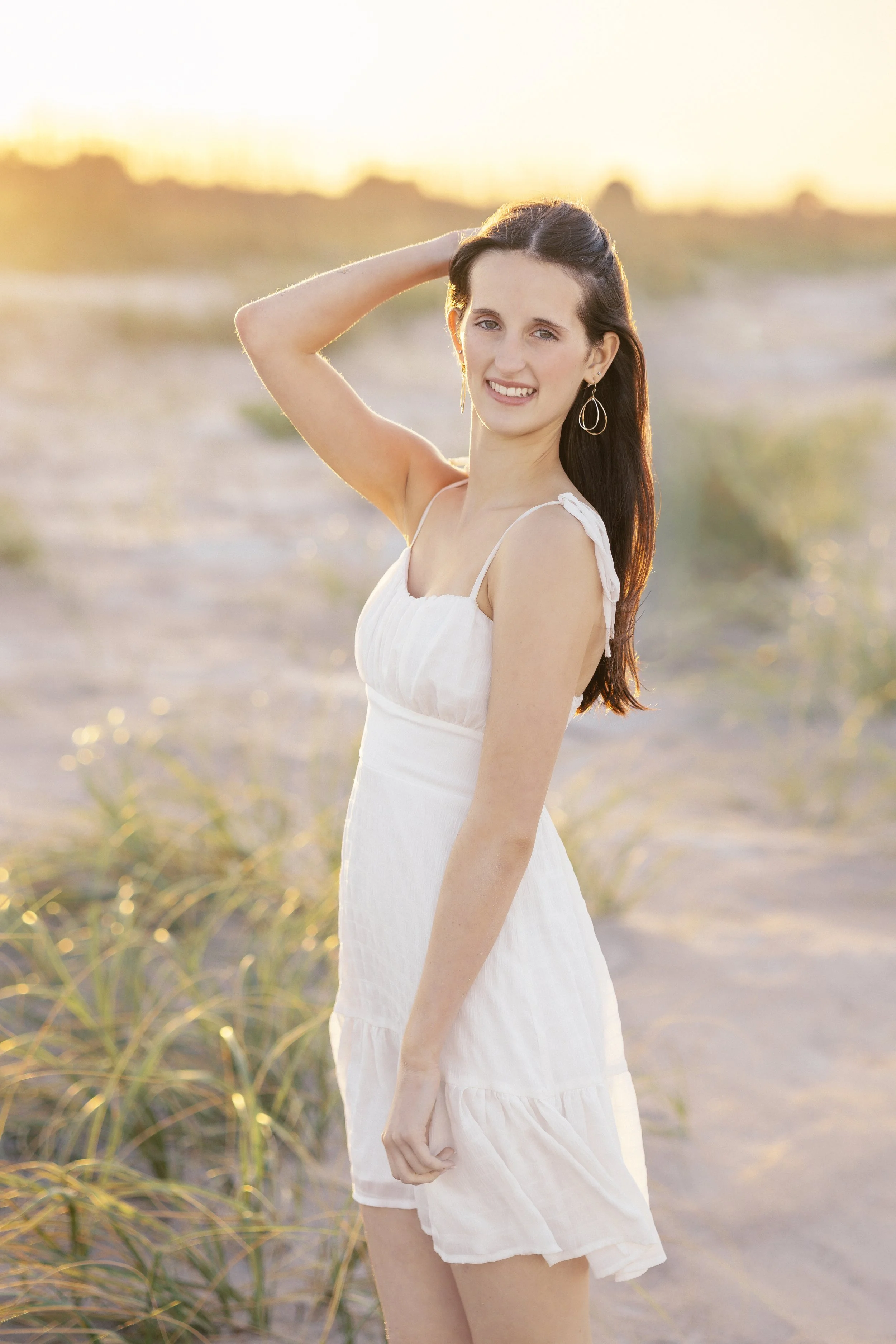 A young woman in a white dress standing outdoors at sunset, with her left hand behind her head and smiling at the camera.