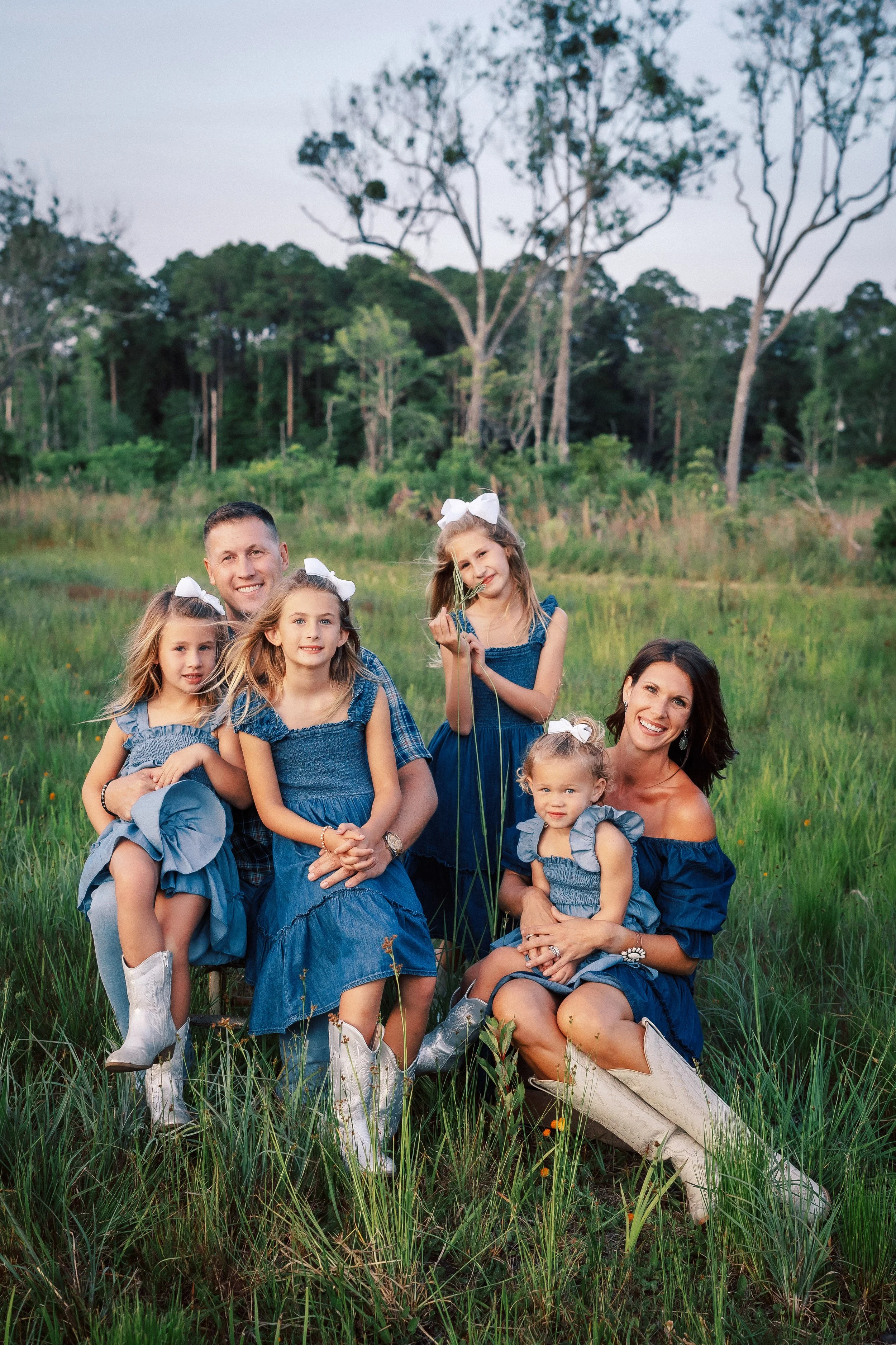 A happy family of seven, including two women, one man, and four young girls, posing in a grassy field with trees in the background during sunset. They are all wearing blue clothing and white boots, and some of the girls have white bows in their hair.