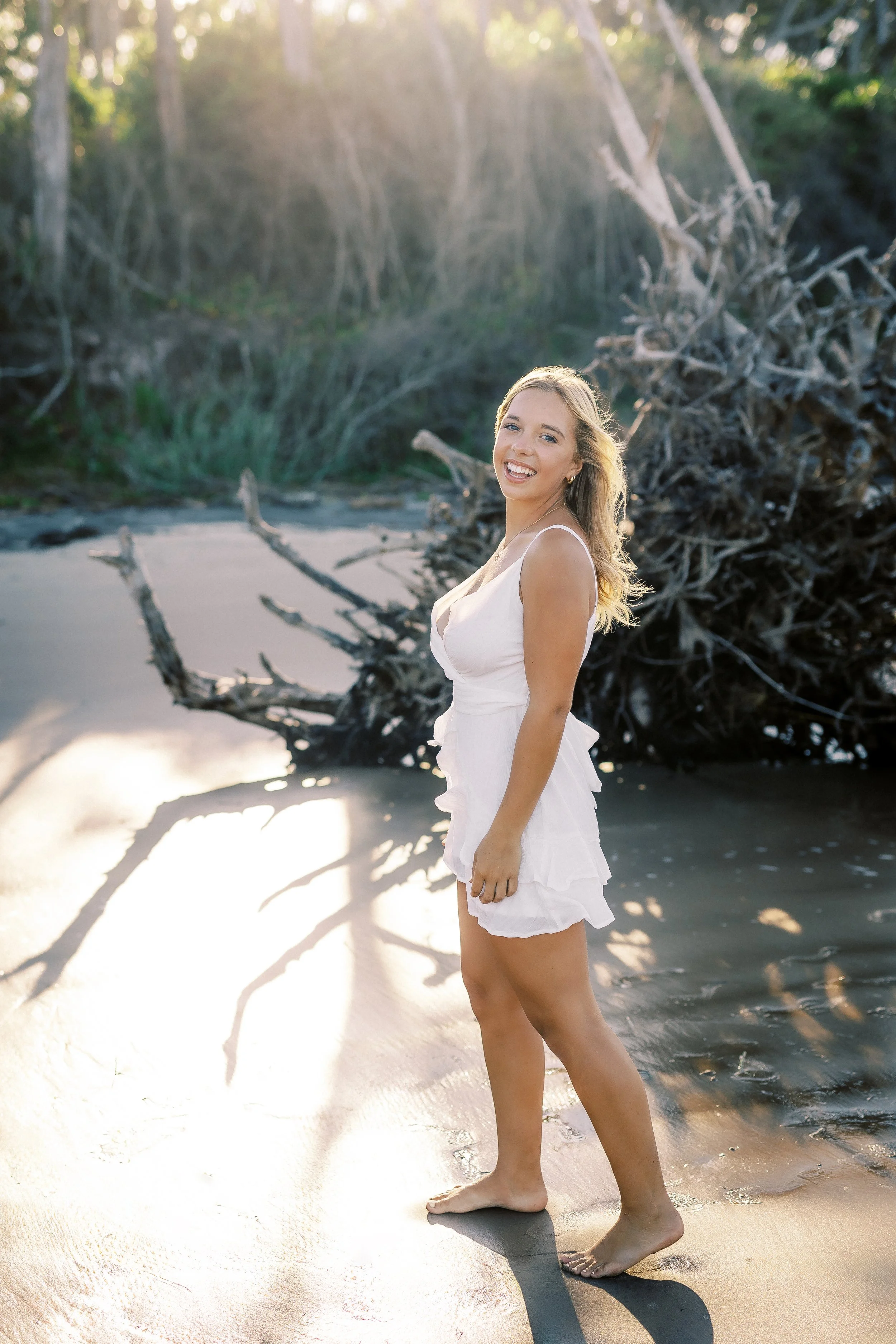 A smiling woman in a white dress standing barefoot on a sandy beach near a driftwood tree at sunset.