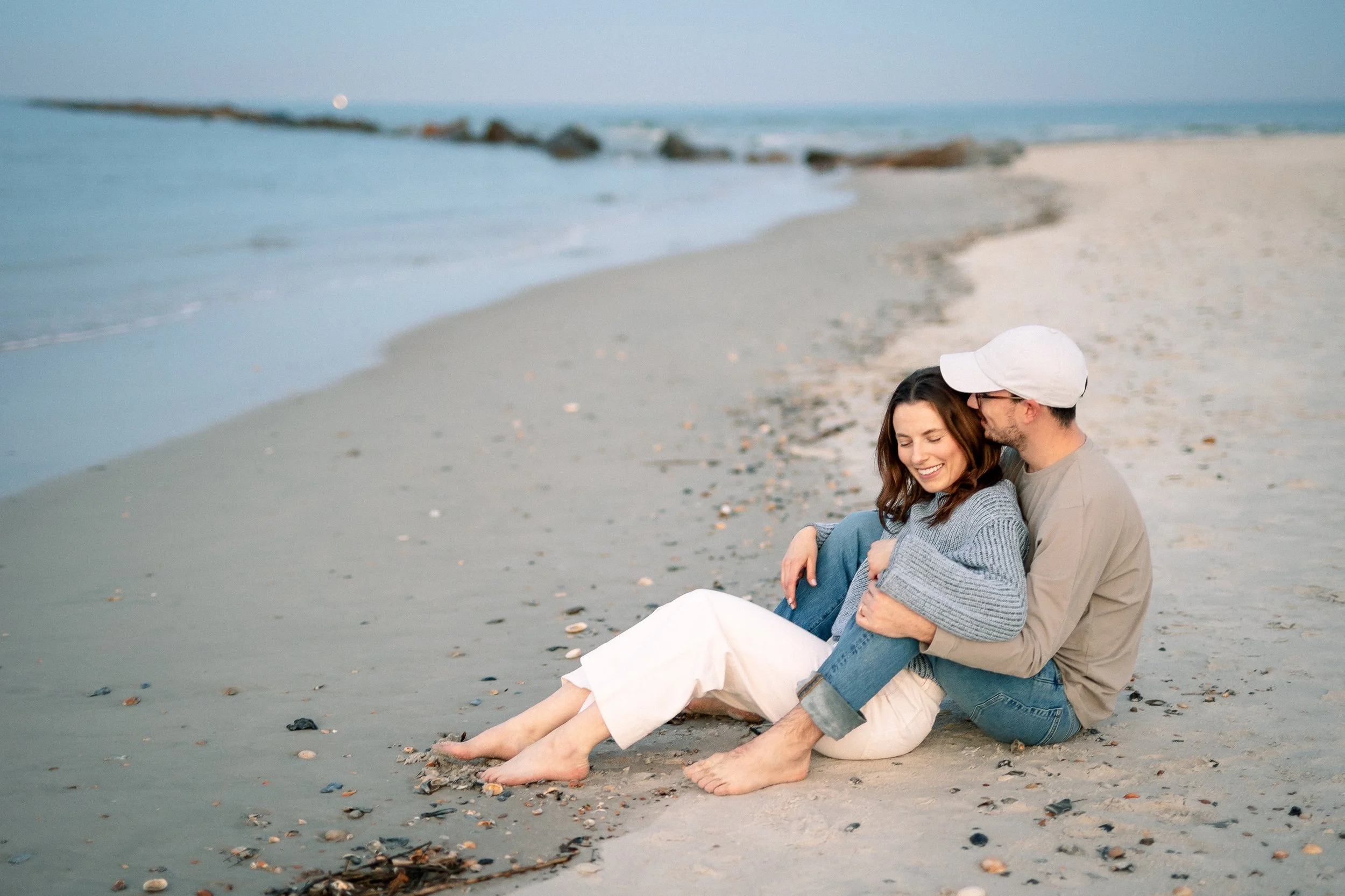 A couple sitting close together on the beach, smiling, with the ocean and rocks in the background.