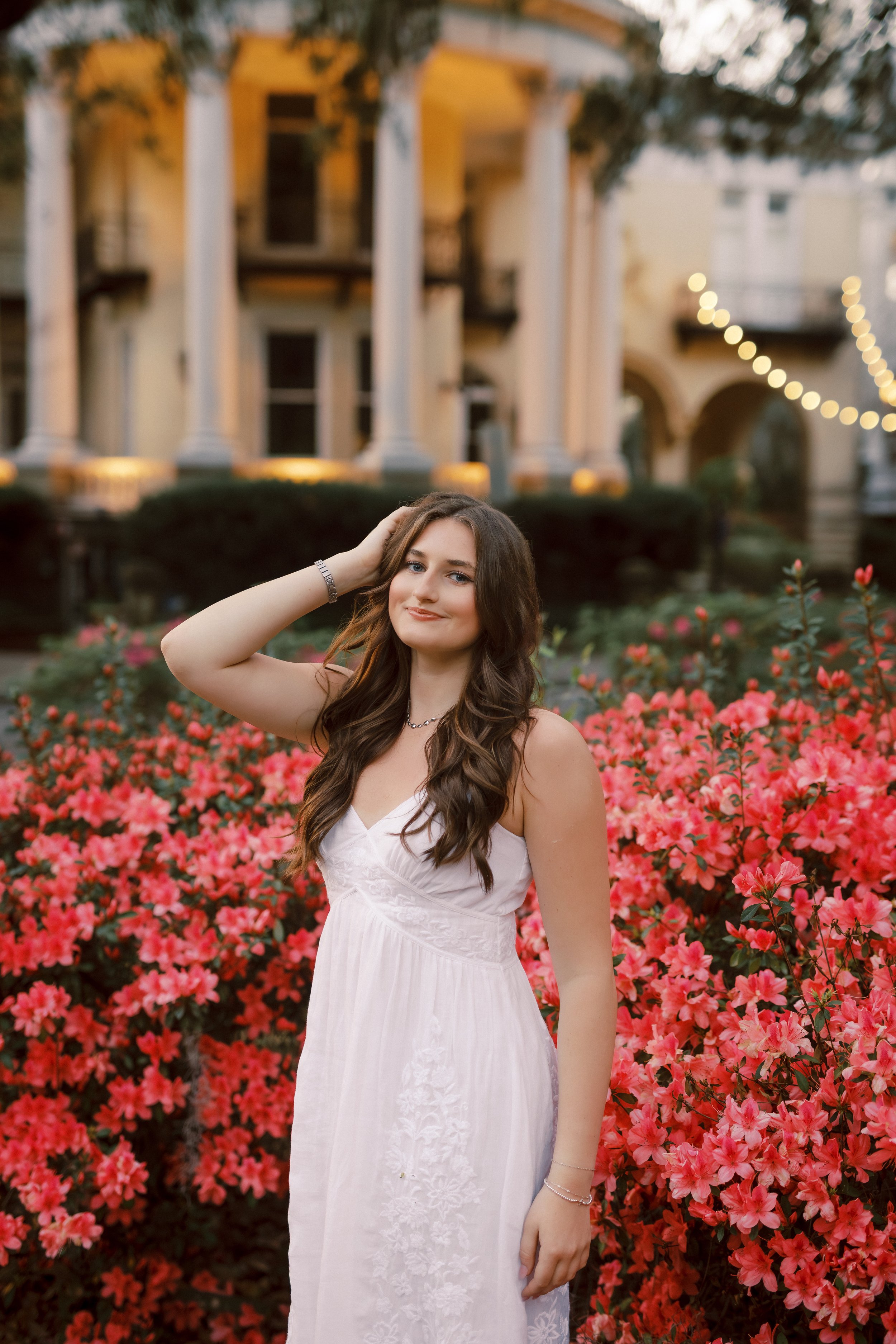 A young woman with long brown hair and light skin wearing a white dress standing among pink azalea flowers in a garden with a large house and string lights in the background.