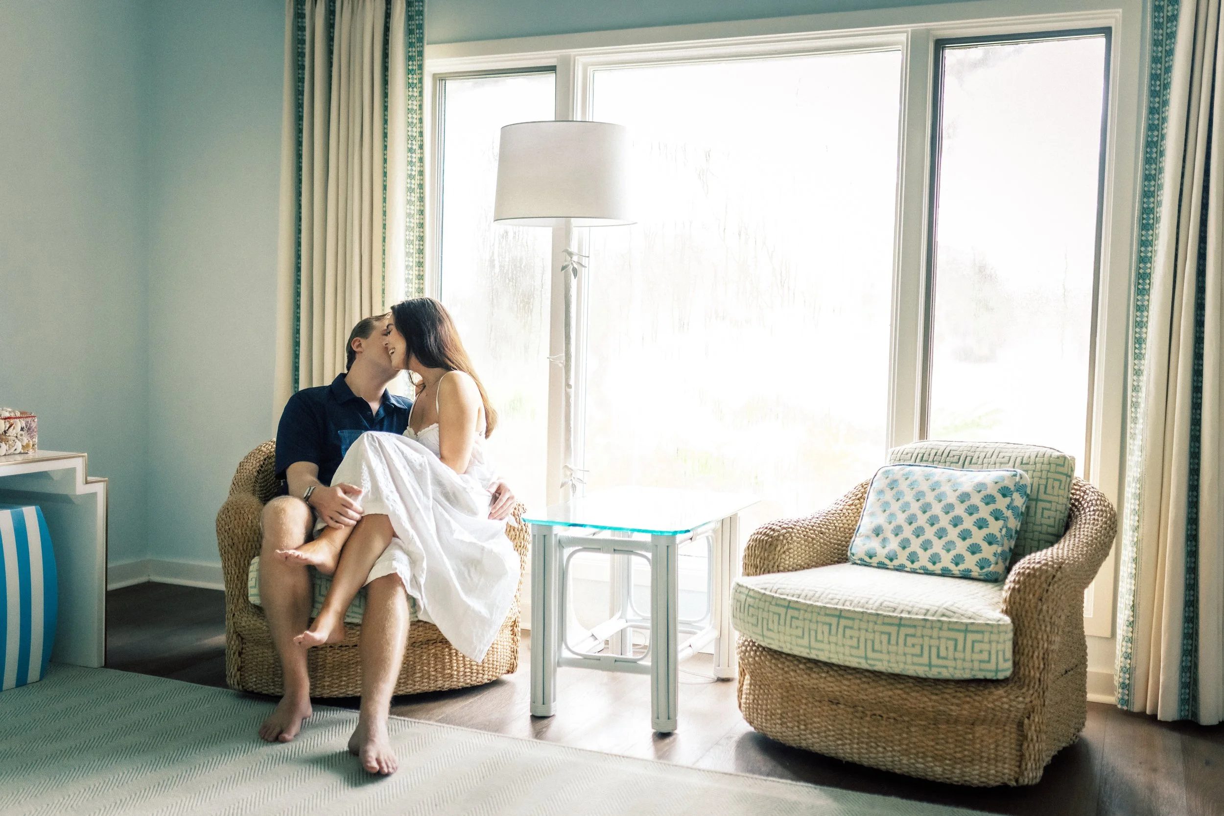 A couple sitting in a bright living room with large windows, the woman sitting on the man's lap and smiling at each other.