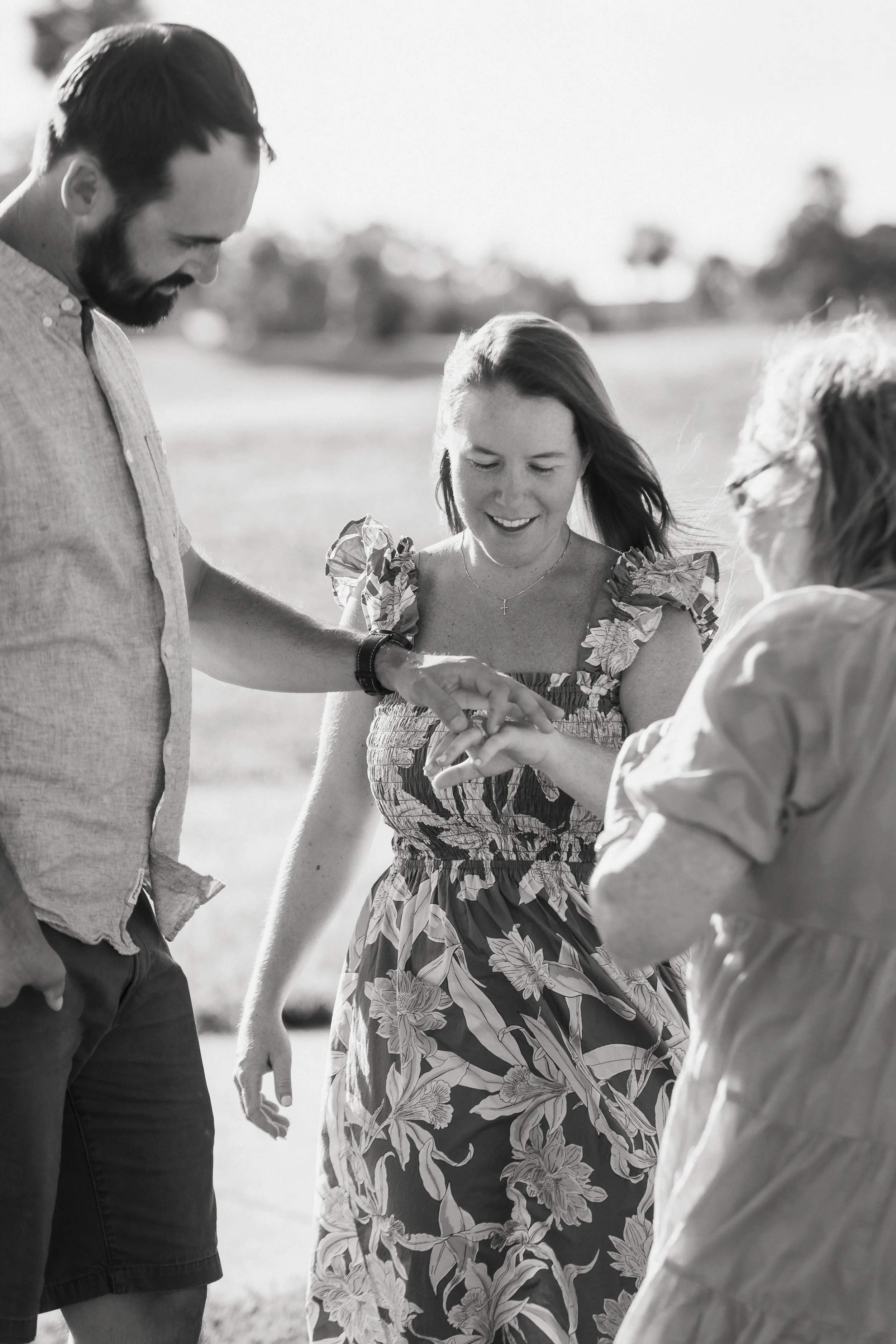 Four people standing outdoors in a circle, holding hands and smiling, with a blurred background of trees and sky, black and white photograph.