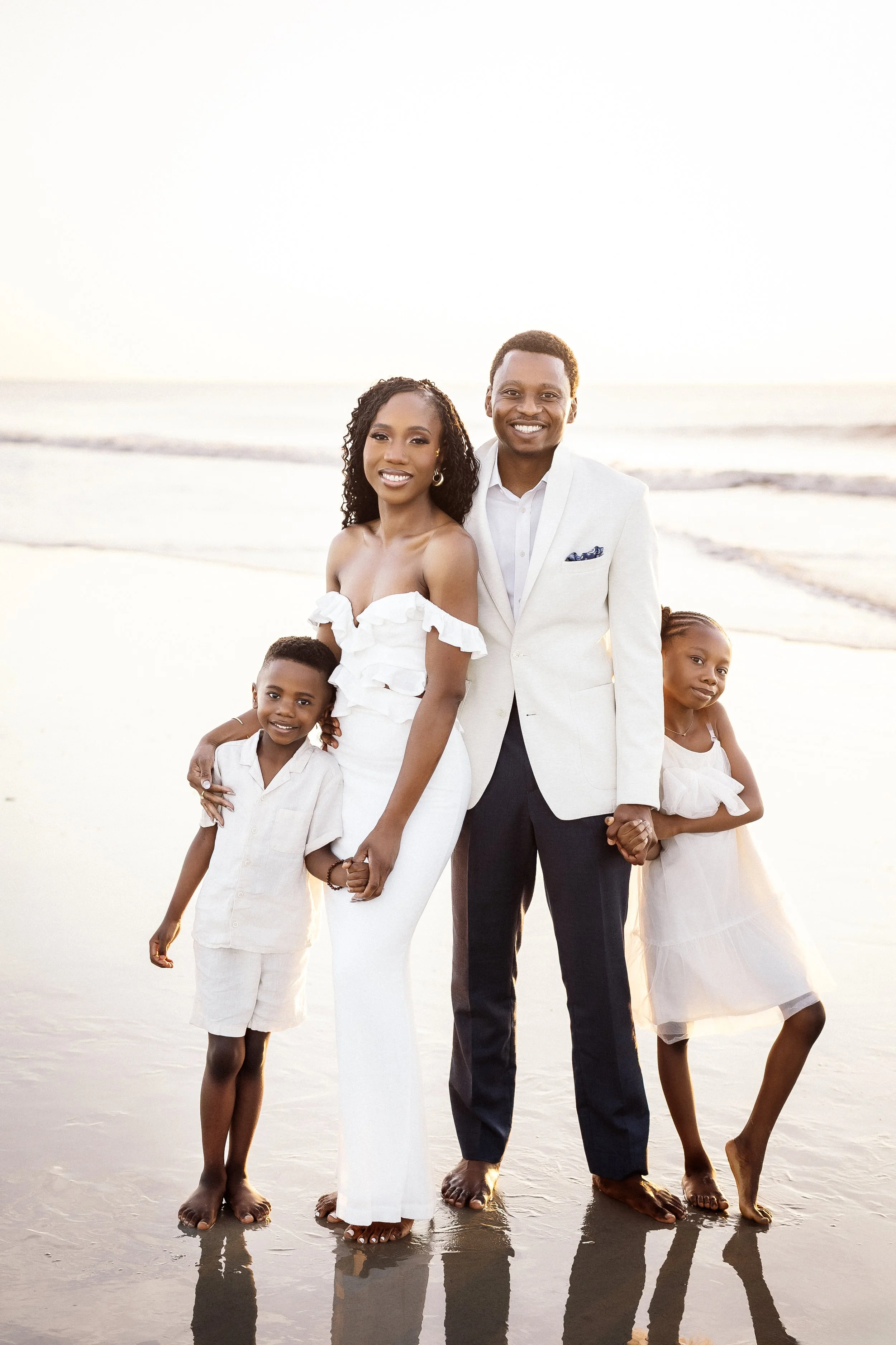 A family of five standing on a beach at sunset, smiling at the camera. The adults are in the center with the woman in a white off-shoulder outfit and the man in a white suit jacket and dark pants. The children, two boys and a girl, are dressed in lig