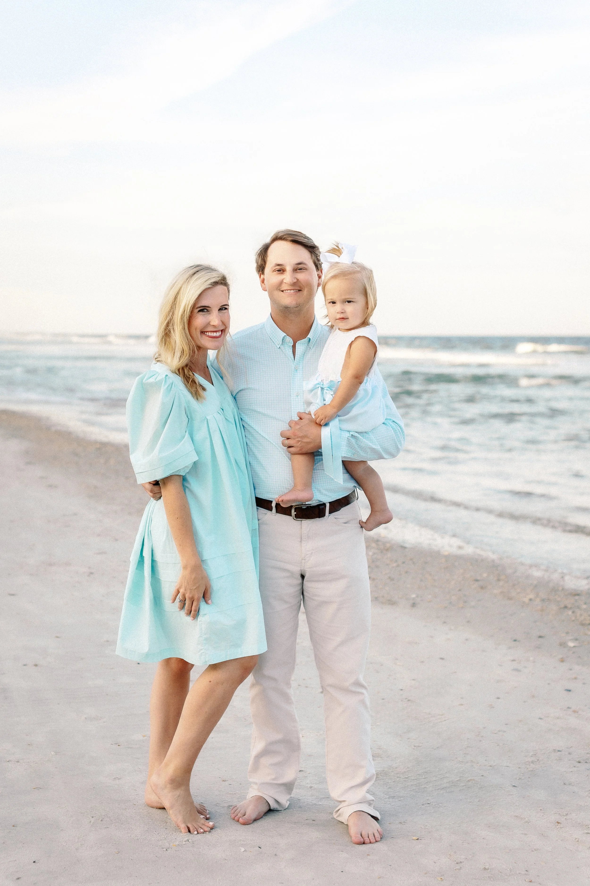 A family of three standing barefoot on the sand at the beach, with ocean waves and a cloudy sky in the background. Photographer Amelia Island, Jacksonville, St Augustine, St Simons Island, Savannah photographer
