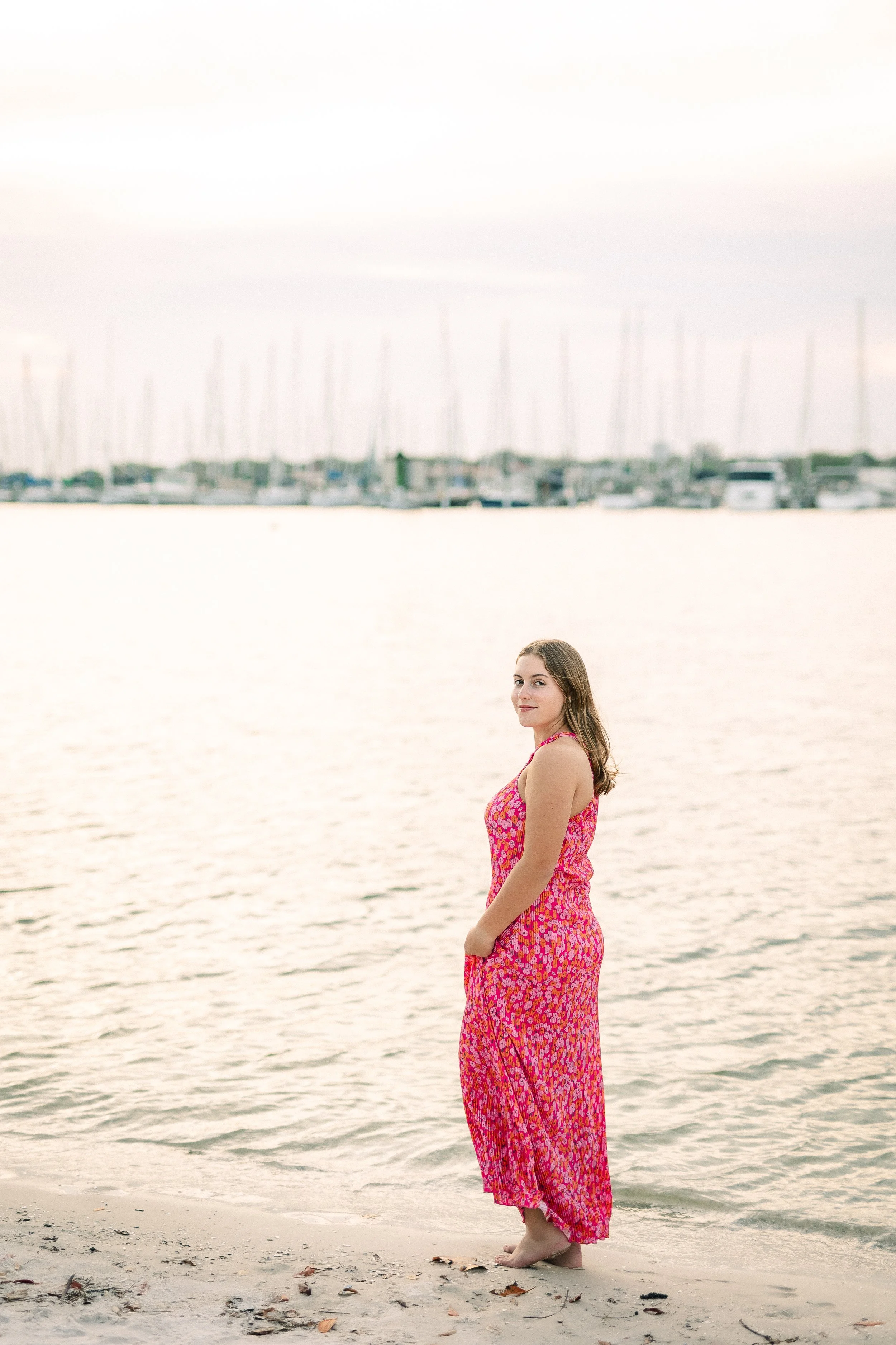 A woman in a pink and orange patterned maxi dress standing barefoot on the beach near the water with sailboats docked in the background.