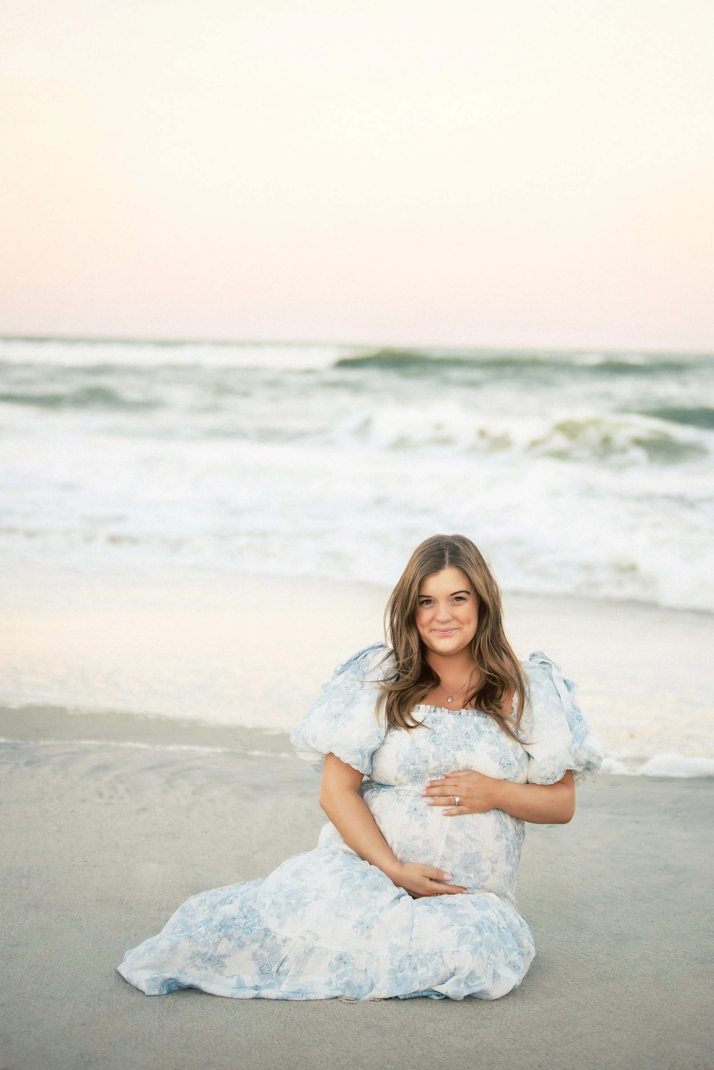 Young woman with long brown hair in a light blue and white floral dress sitting on the beach with ocean waves in the background