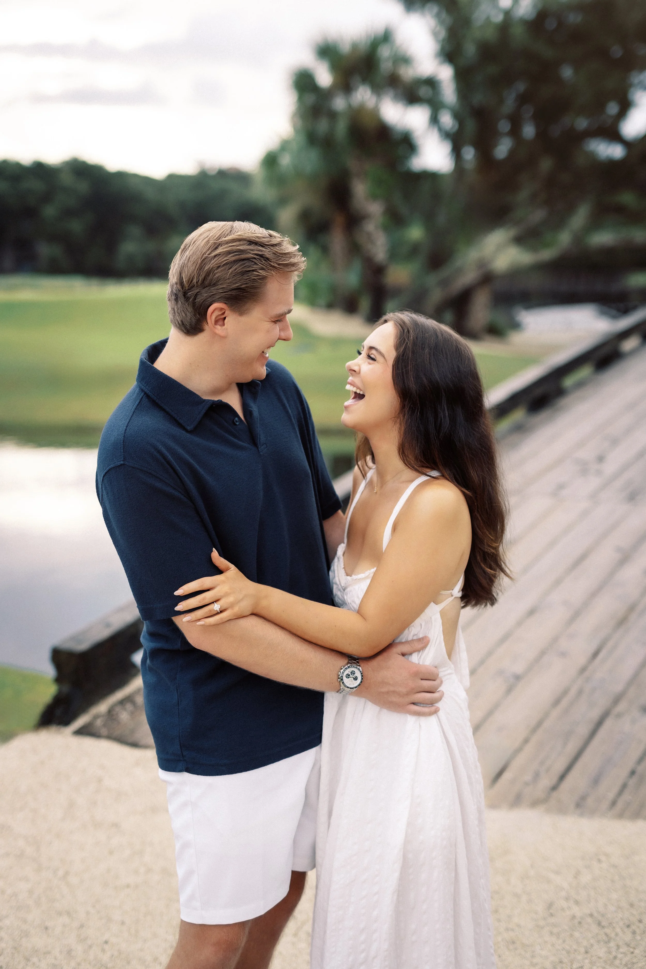 A smiling couple embracing outdoors near a lake or river with trees in the background, a wooden pier, and overcast sky.