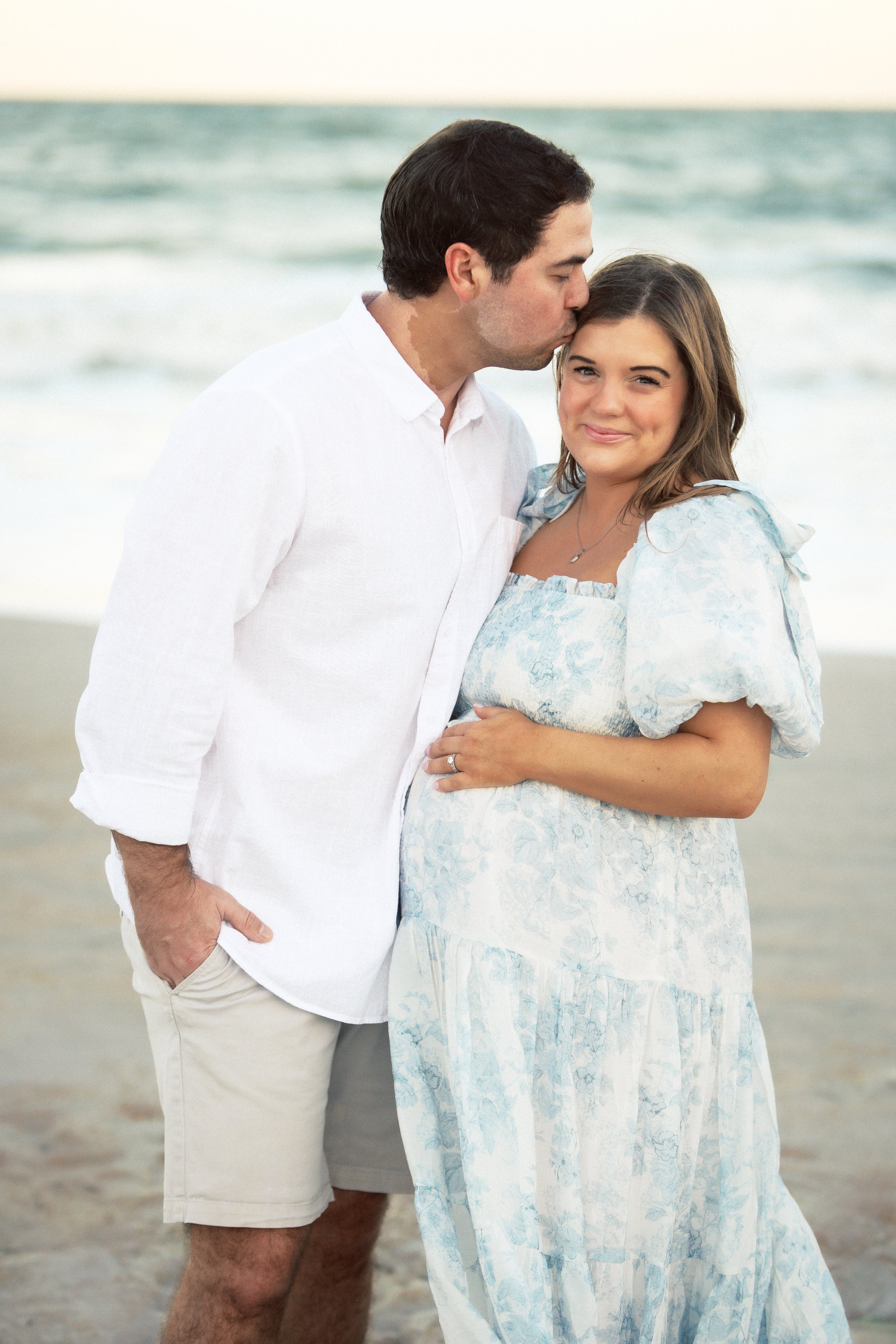 A man and woman standing on a beach, with the man kissing the woman's forehead. The woman is smiling and looking at the camera. The ocean is in the background.